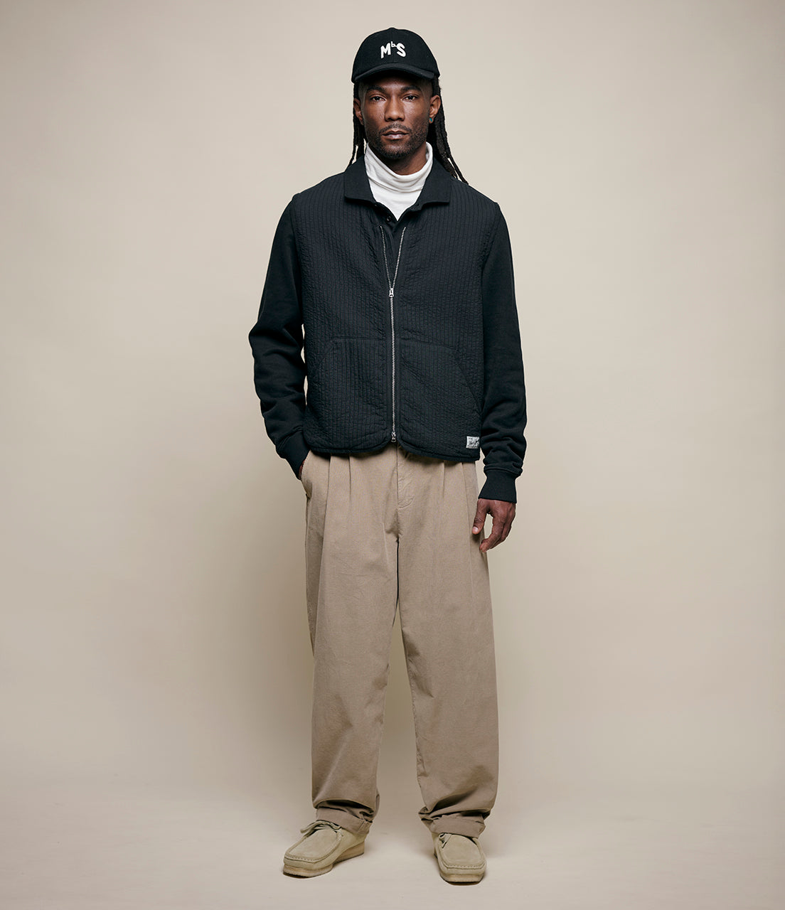 Man wearing a black vest, beige pants, and a black cap on a beige background