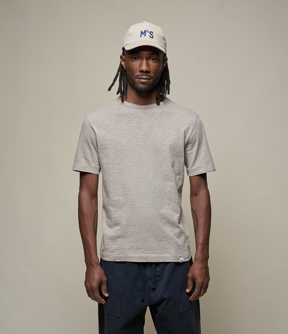 Man wearing a gray t-shirt and white cap with 'MbS' logo on a beige background