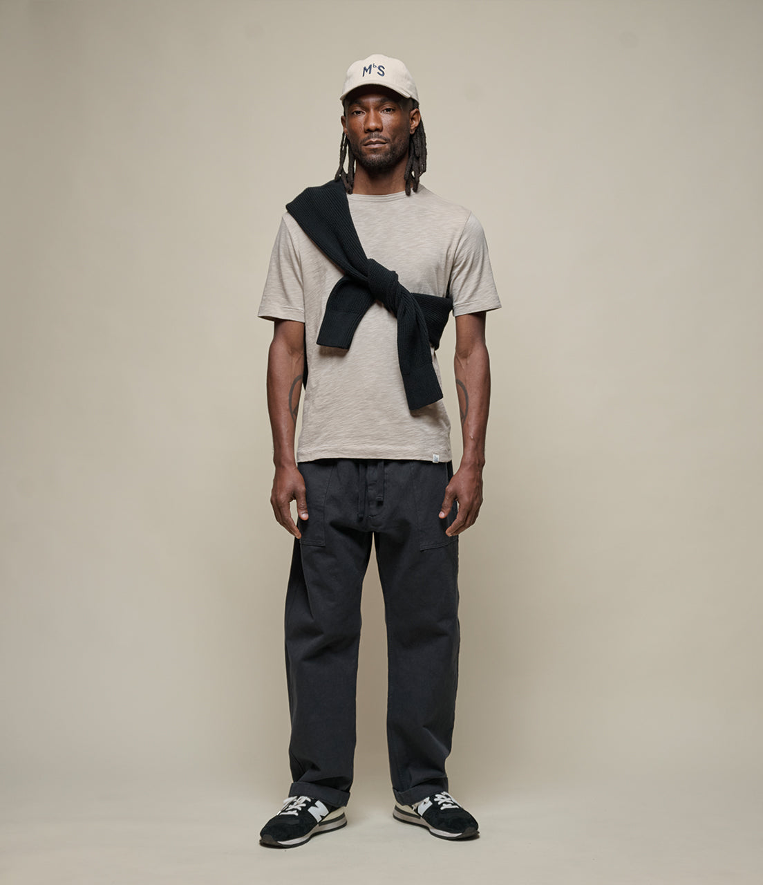 Man wearing a beige t-shirt, dark pants, and a cap with a scarf over his shoulder on a plain background