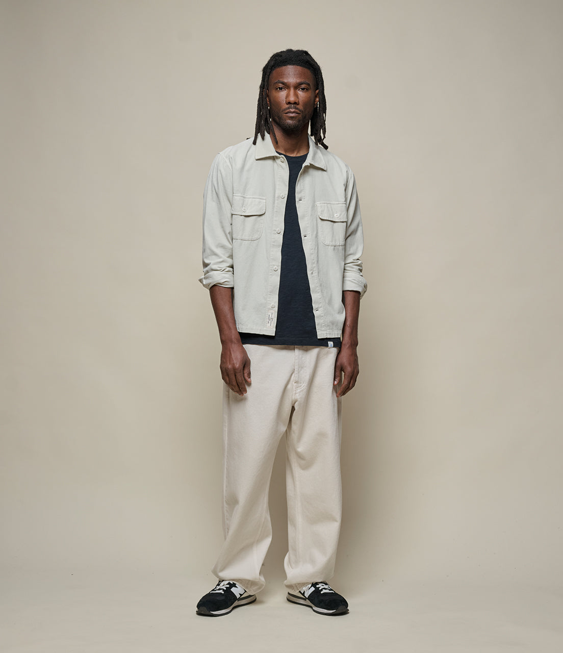 Man wearing a light twill shirt, dark t-shirt, white pants, and black sneakers on a beige background