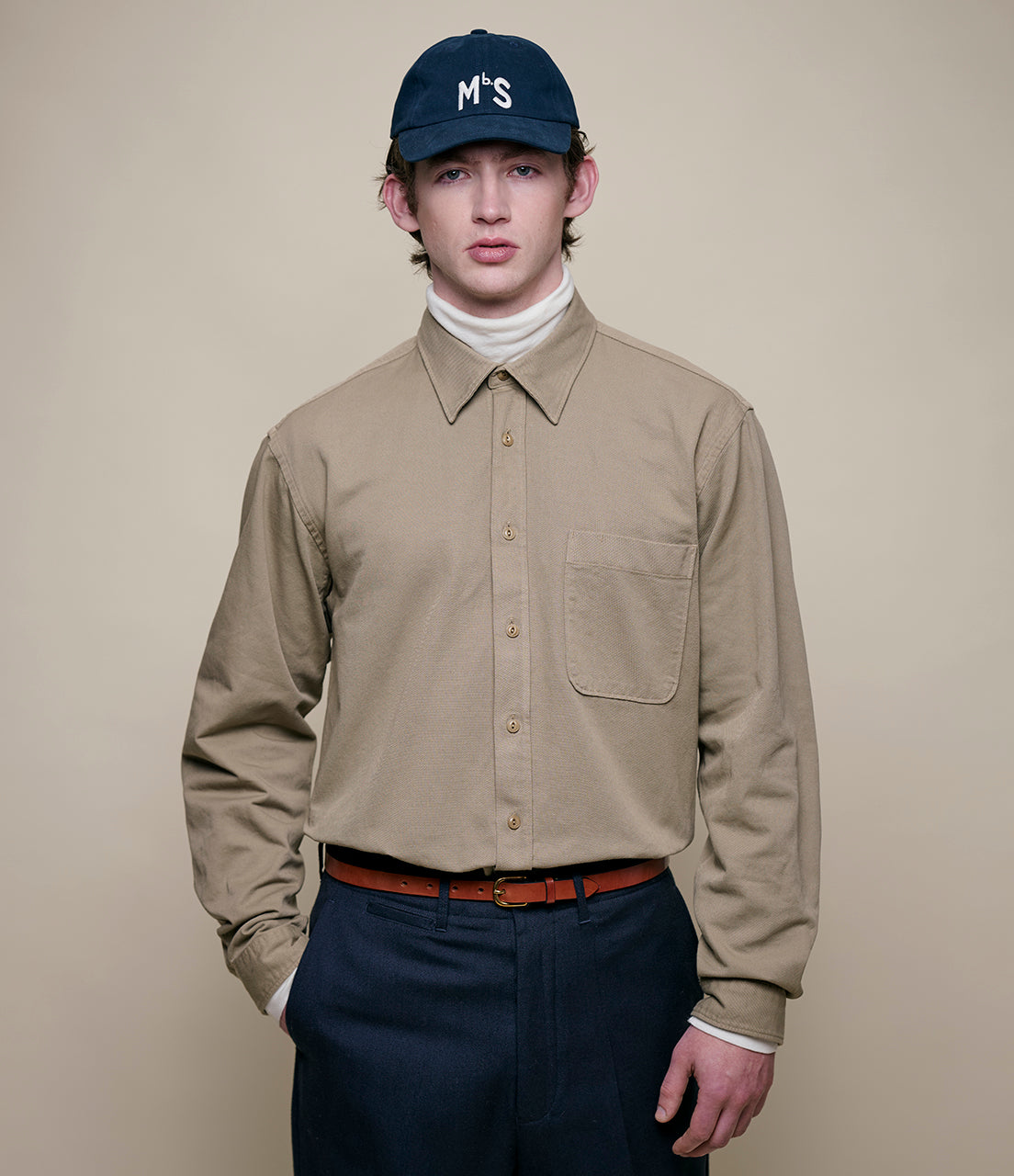 Man wearing a beige shirt, navy pants, and a navy cap with 'M'S' logo on a beige background