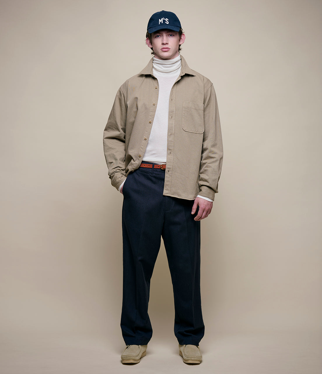 Person wearing a beige jacket, navy pants, and a navy cap on a plain background