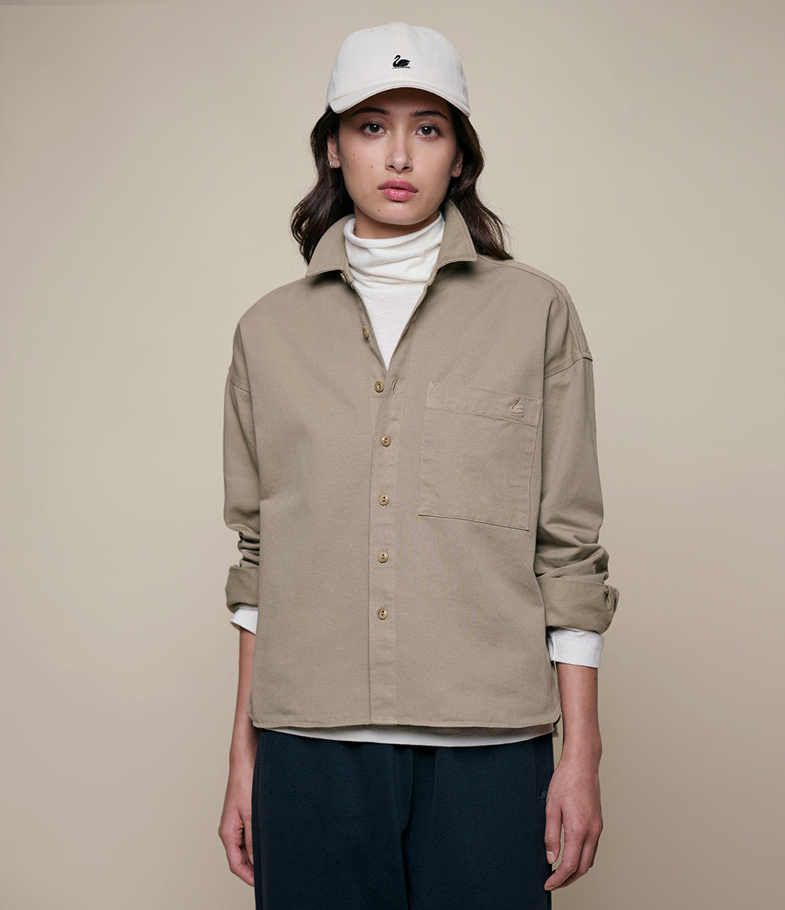 Person wearing a beige twill shirt, white turtleneck, and black pants on a beige background