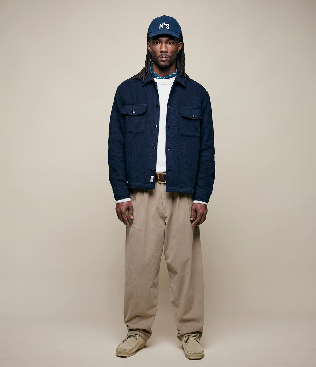 Person wearing a navy jacket, white shirt, beige pants, and a navy cap on a beige background
