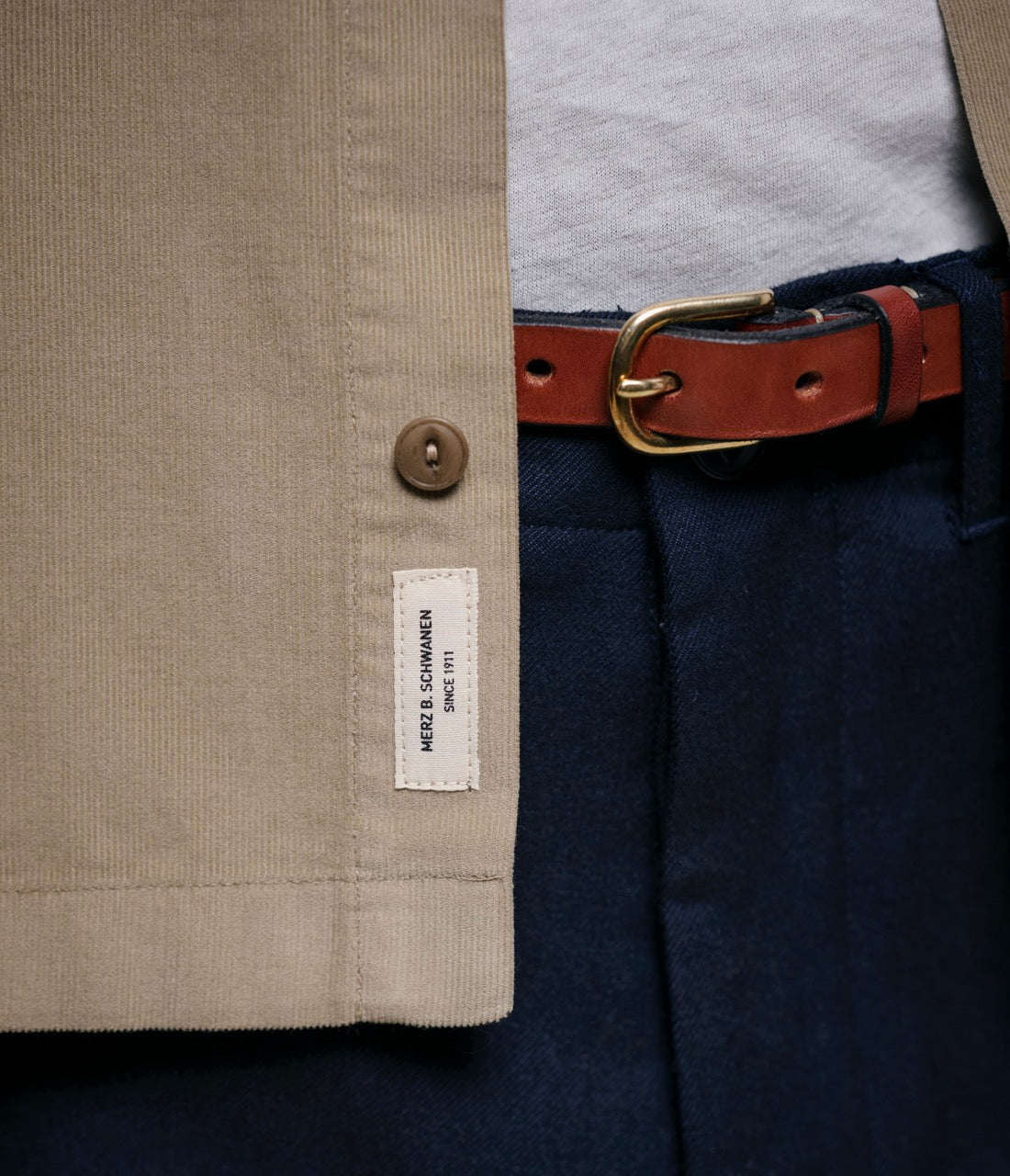 Close-up of a beige shirt with a label, navy pants, and brown belt.