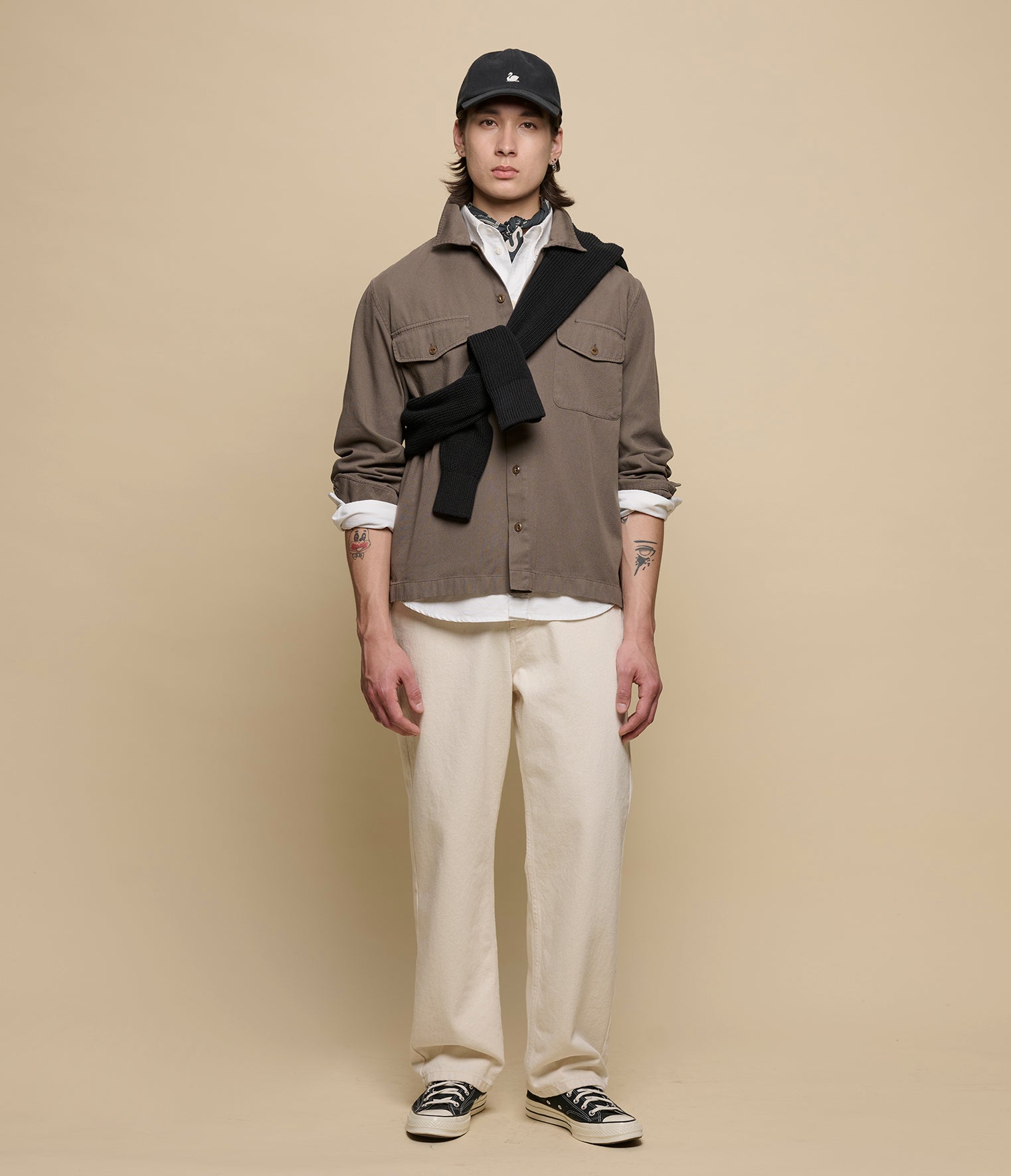Person wearing a brown jacket, white pants, and black cap against a beige background