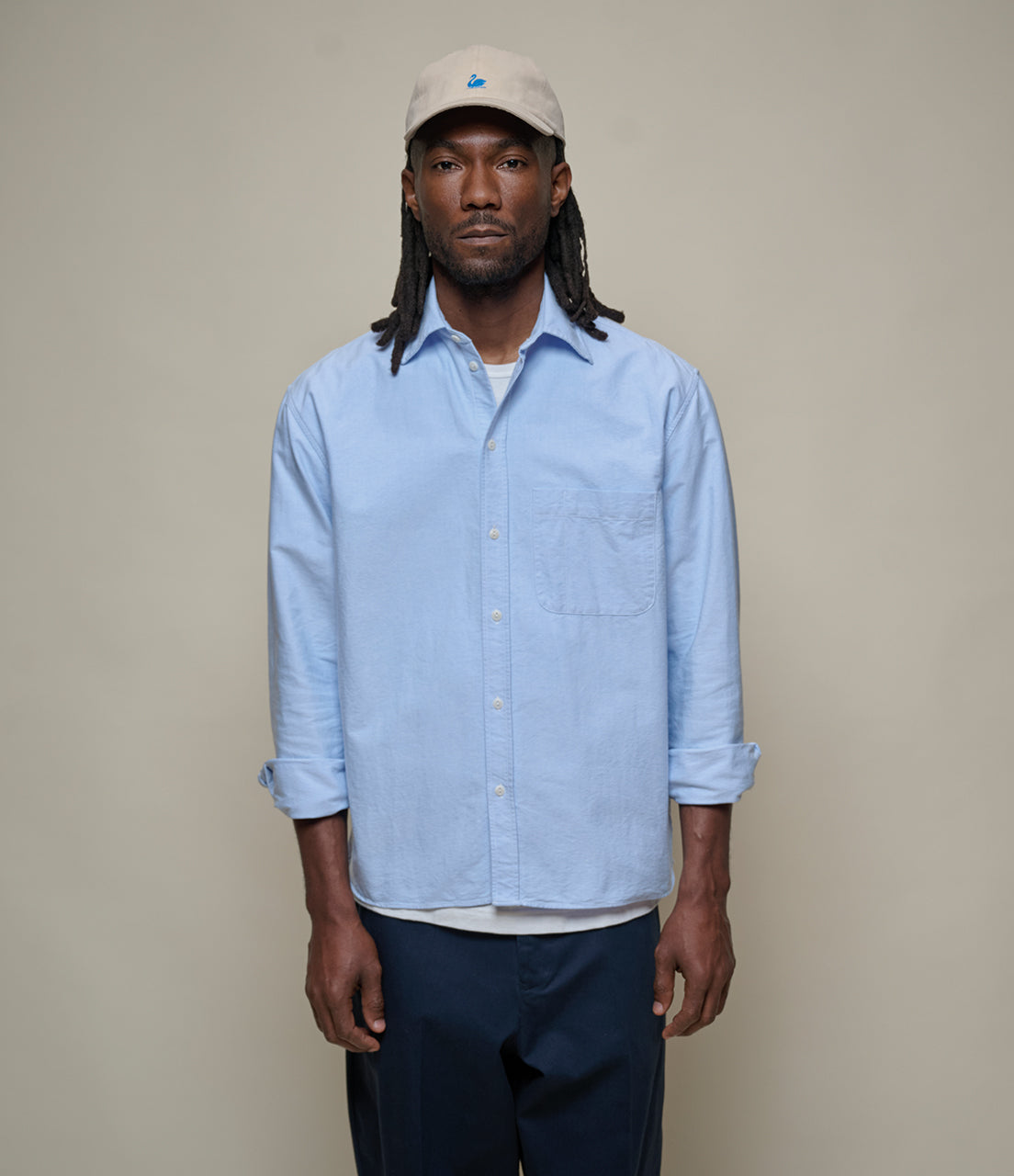 Man wearing a light blue oxford shirt and beige cap against a plain background