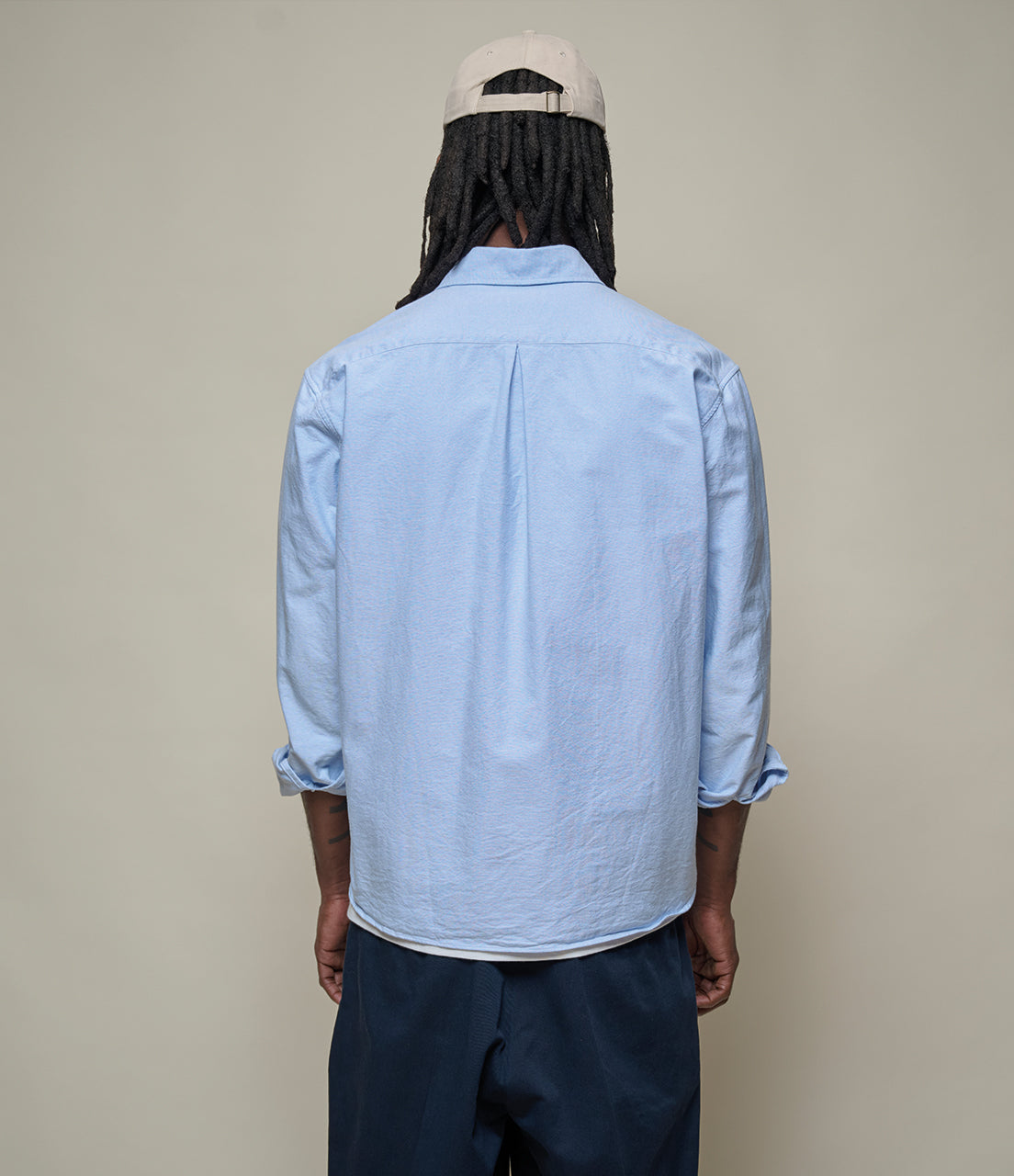 Person wearing a light blue shirt and dark pants with a beige cap on a plain background