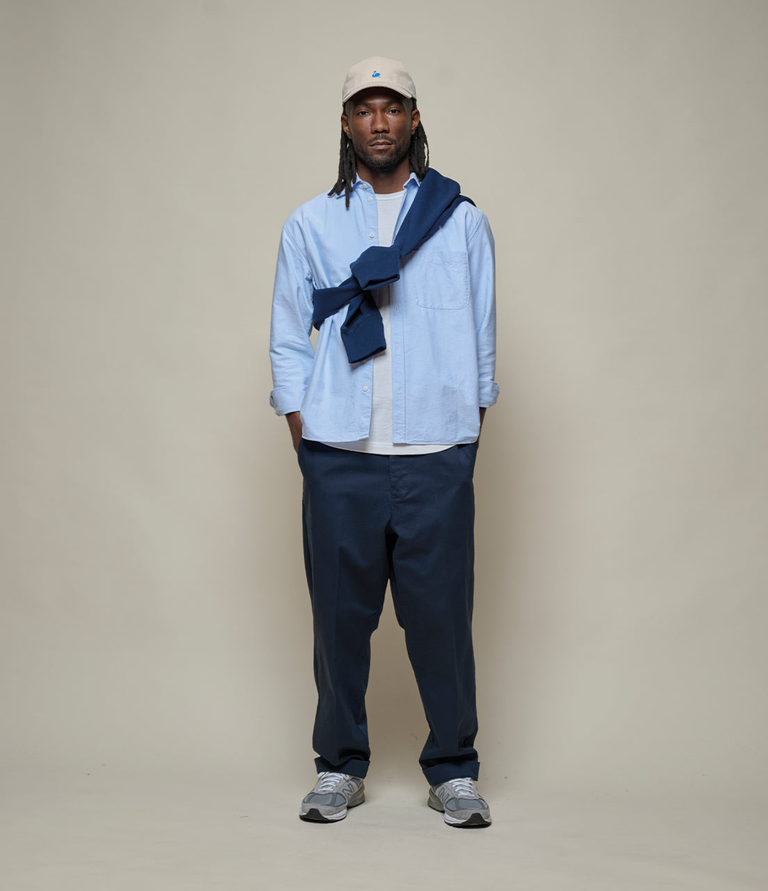 Person wearing a light blue shirt, dark pants, and a beige cap against a plain background