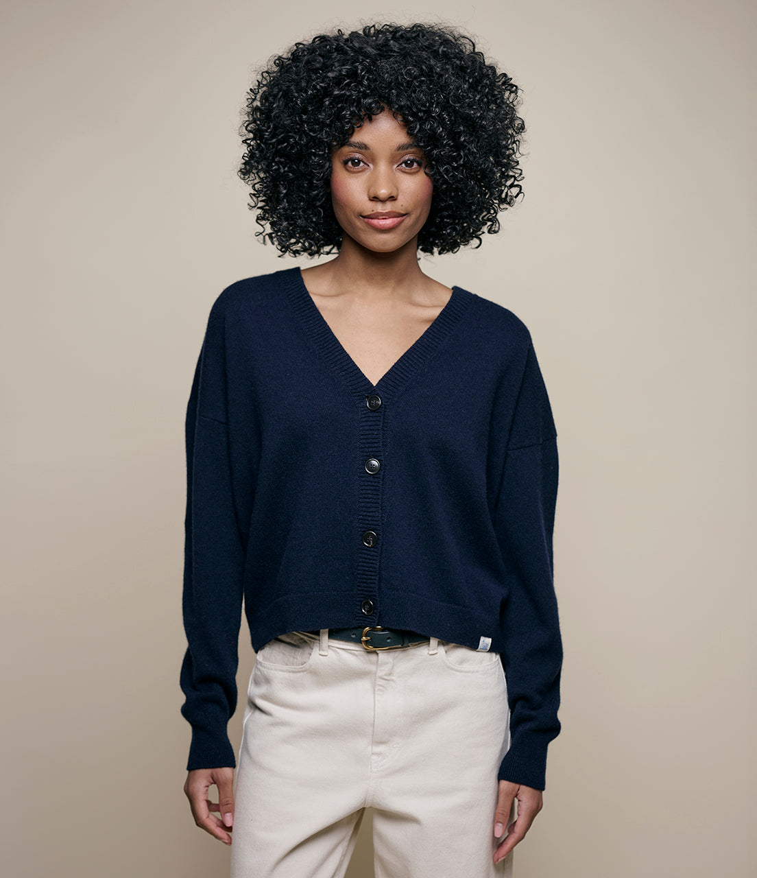 A person with curly hair wearing a navy blue cropped cardigan with large buttons and a V-neckline, paired with white high-waisted jeans.
