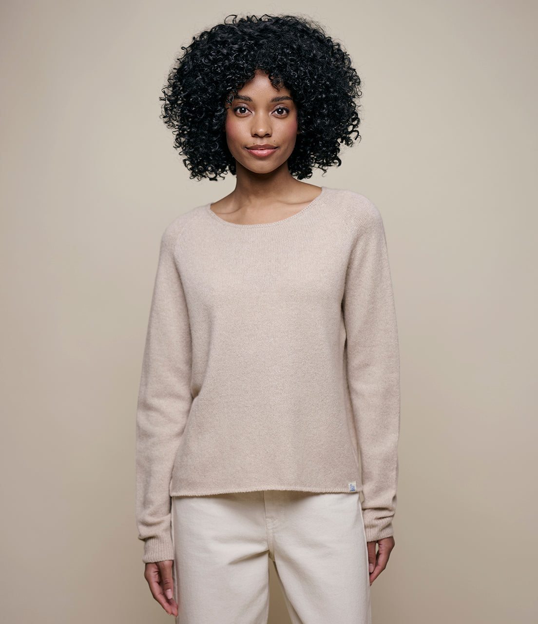 A person with curly hair wearing a beige long-sleeved sweater with a round neckline.