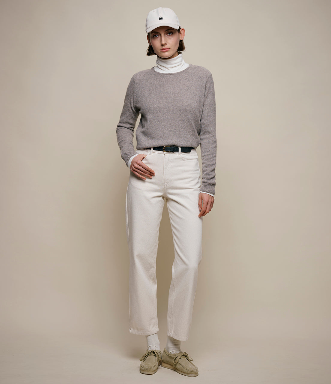 Person wearing a gray sweater, white pants, and a white cap on a beige background