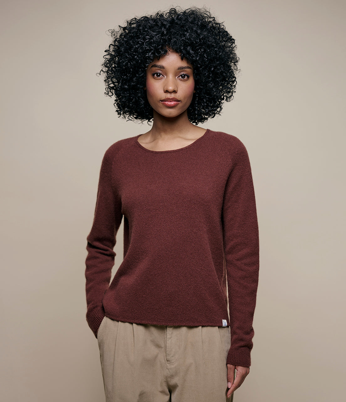 A person with curly hair wearing a brown long-sleeved, round-neck Merino Wool-Cashmere-Silk Raglan Sweater paired with light-colored pants.