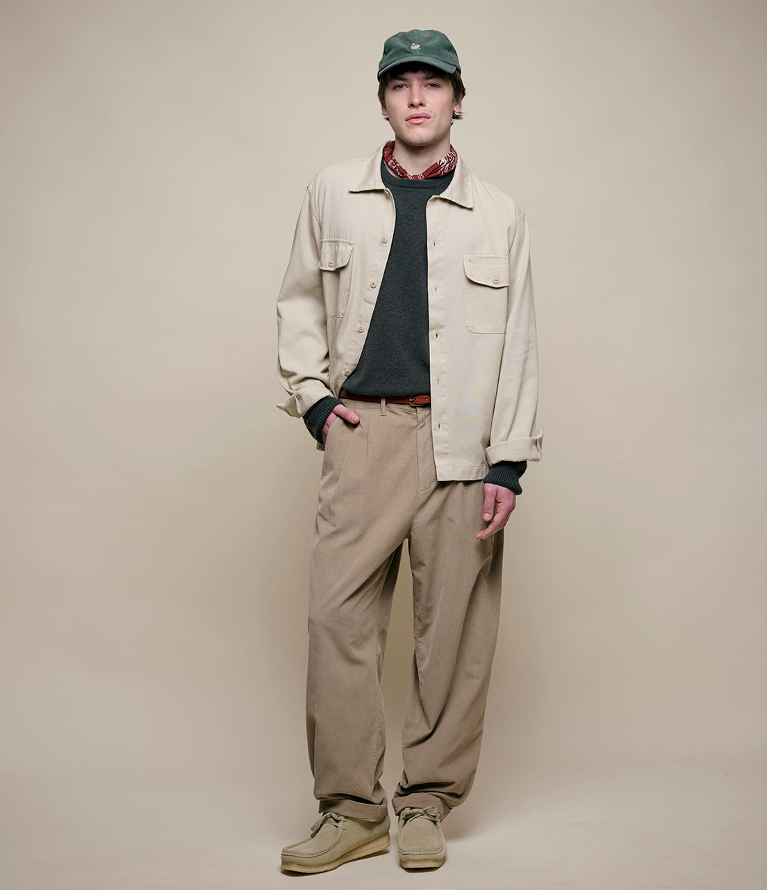 Man wearing a beige jacket, khaki pants, and a green cap against a plain background