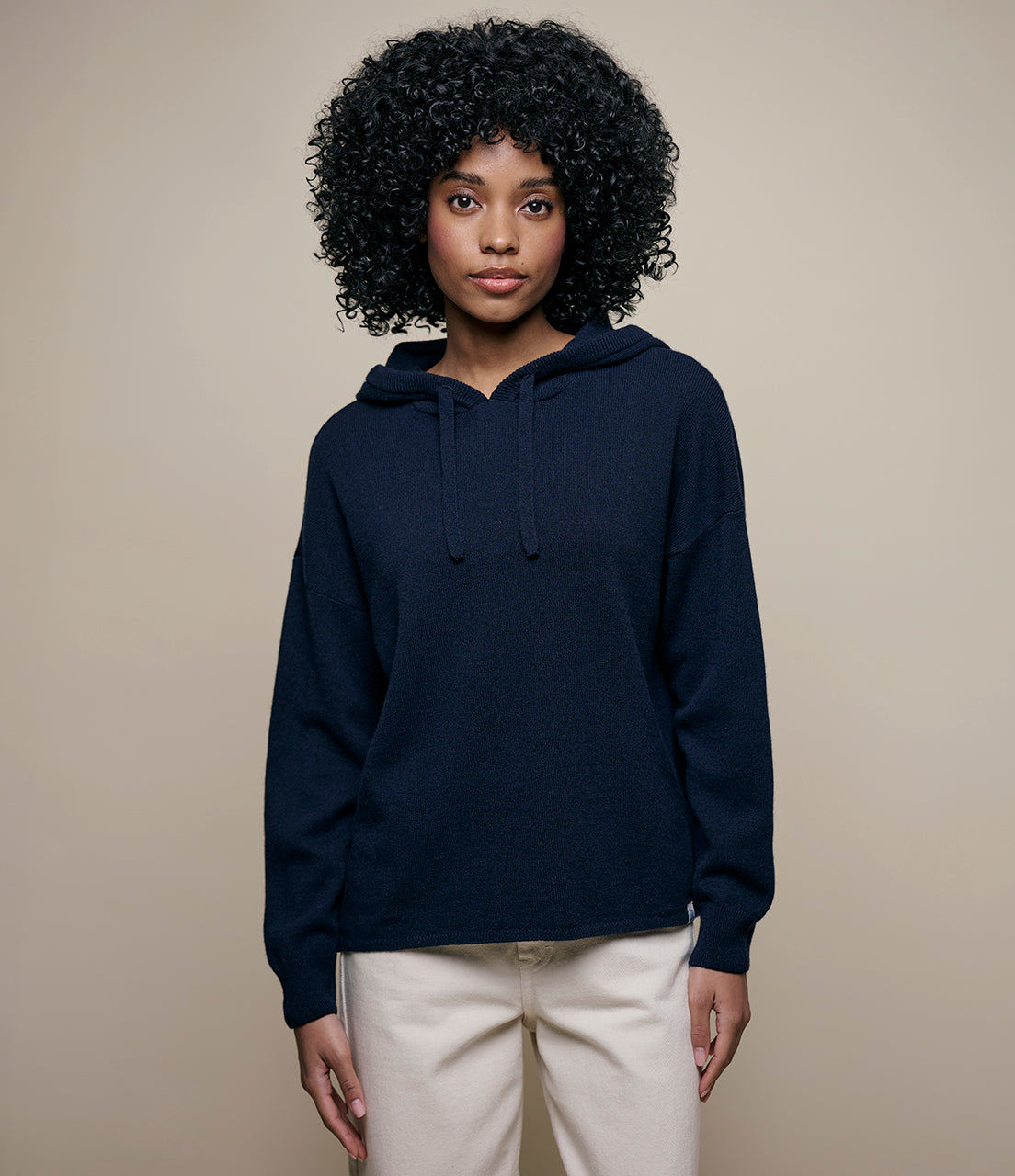A woman wearing a navy blue hoodie with the hood on and light-colored pants. She has curly hair and is posing against a neutral background.
