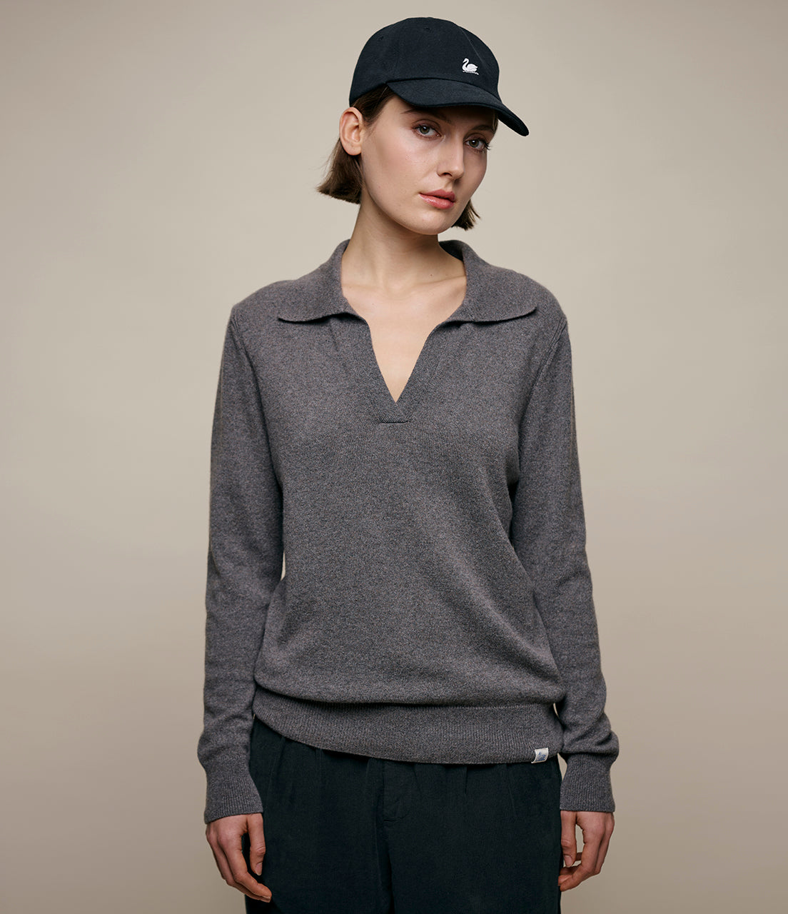 A person wearing a gray V-neck sweater with long sleeves, paired with black pants and a black cap.