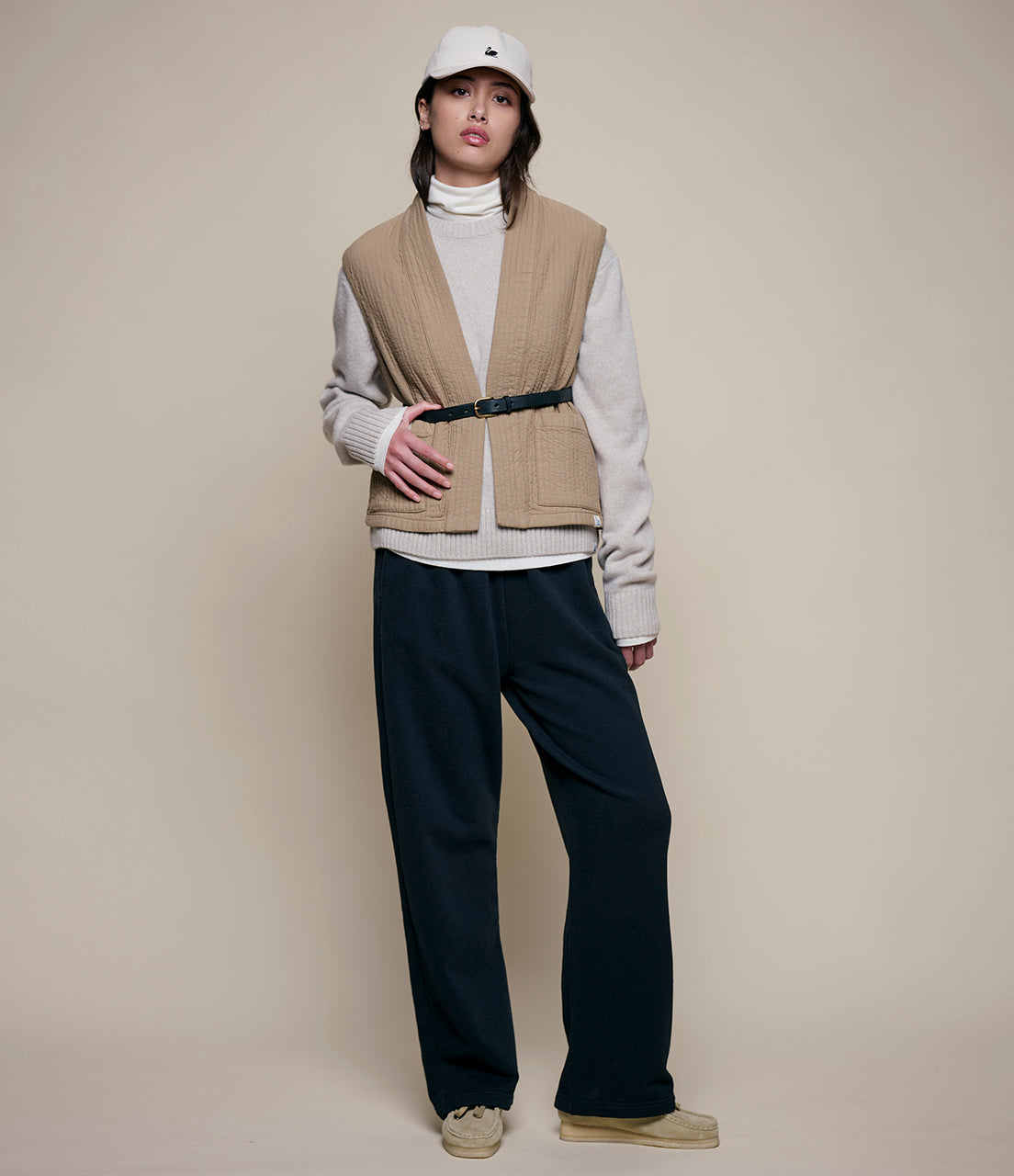 Person wearing a beige vest, white sweater, black pants, and a white cap on a beige background