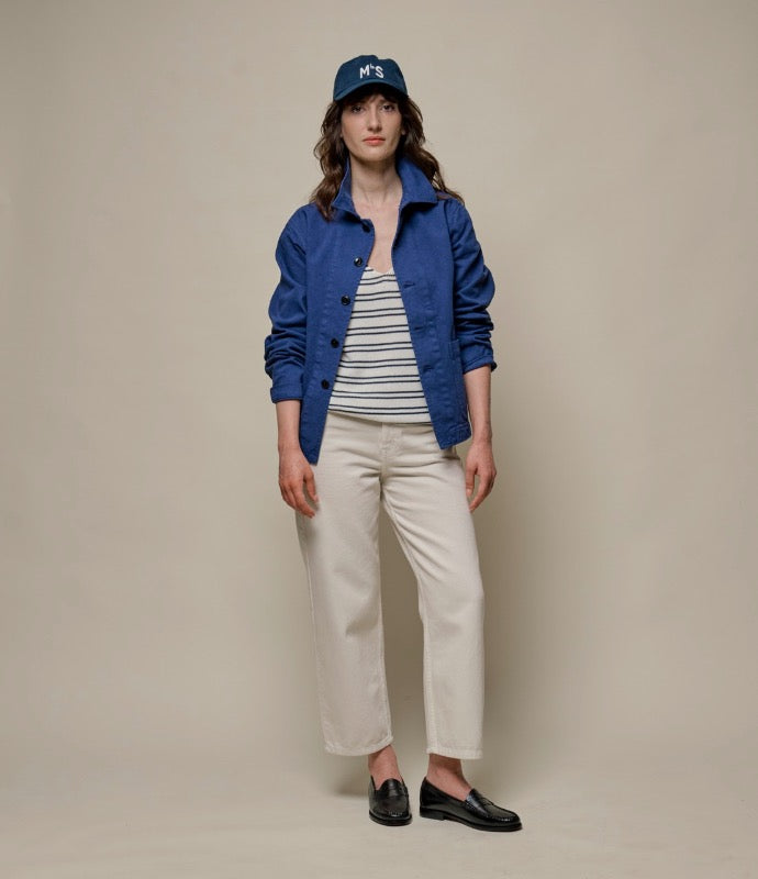 A model wearing a blue cropped twill jacket with a striped shirt, white trousers, and black shoes, also sporting a cap.