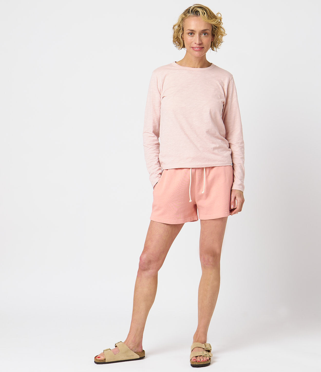 A woman standing against a white background wearing a pink longsleeve shirt and matching pink shorts, with beige sandals and beige Accessories.