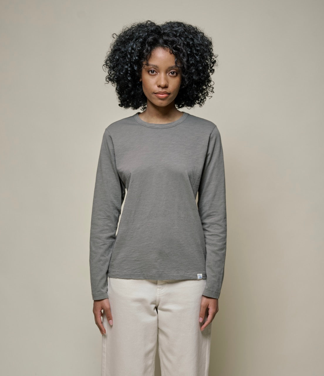 A woman wearing a long-sleeved gray t-shirt and white pants standing against a beige background.