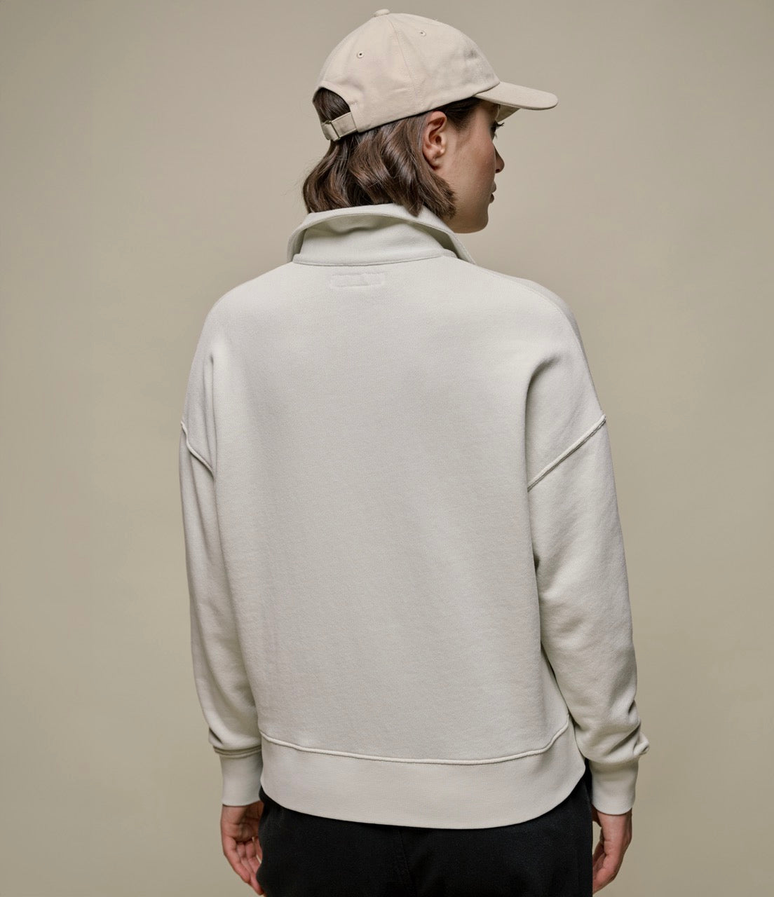 Backside of a woman wearing a beige half-zip sweatshirt with a stand-up collar and black pants.