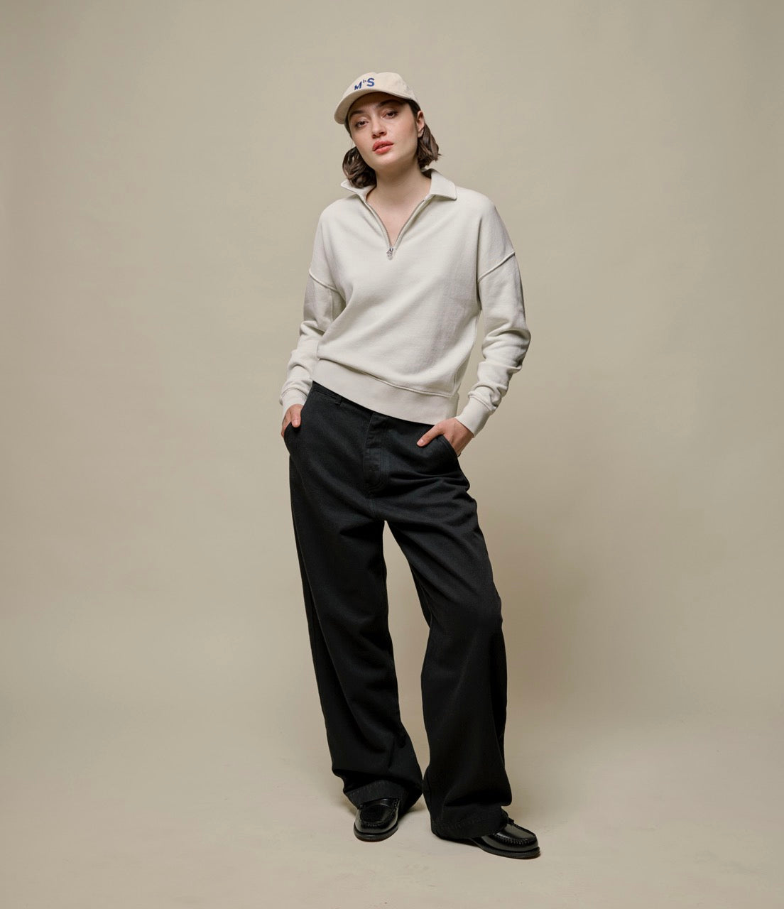 A woman wearing a beige half-zip sweatshirt with a stand-up collar and black pants. She has a cap on her head and is posing with her hands in her pockets.