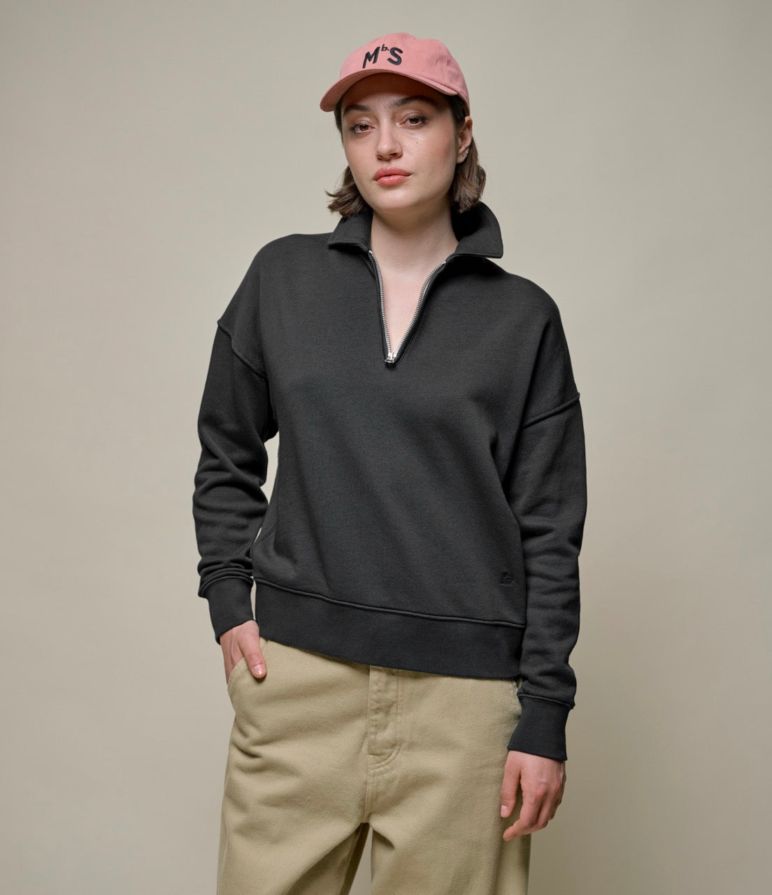 A woman wearing a black half-zip sweatshirt with a stand-up collar, beige pants, and a pink cap with the letters 'MbS' on it.