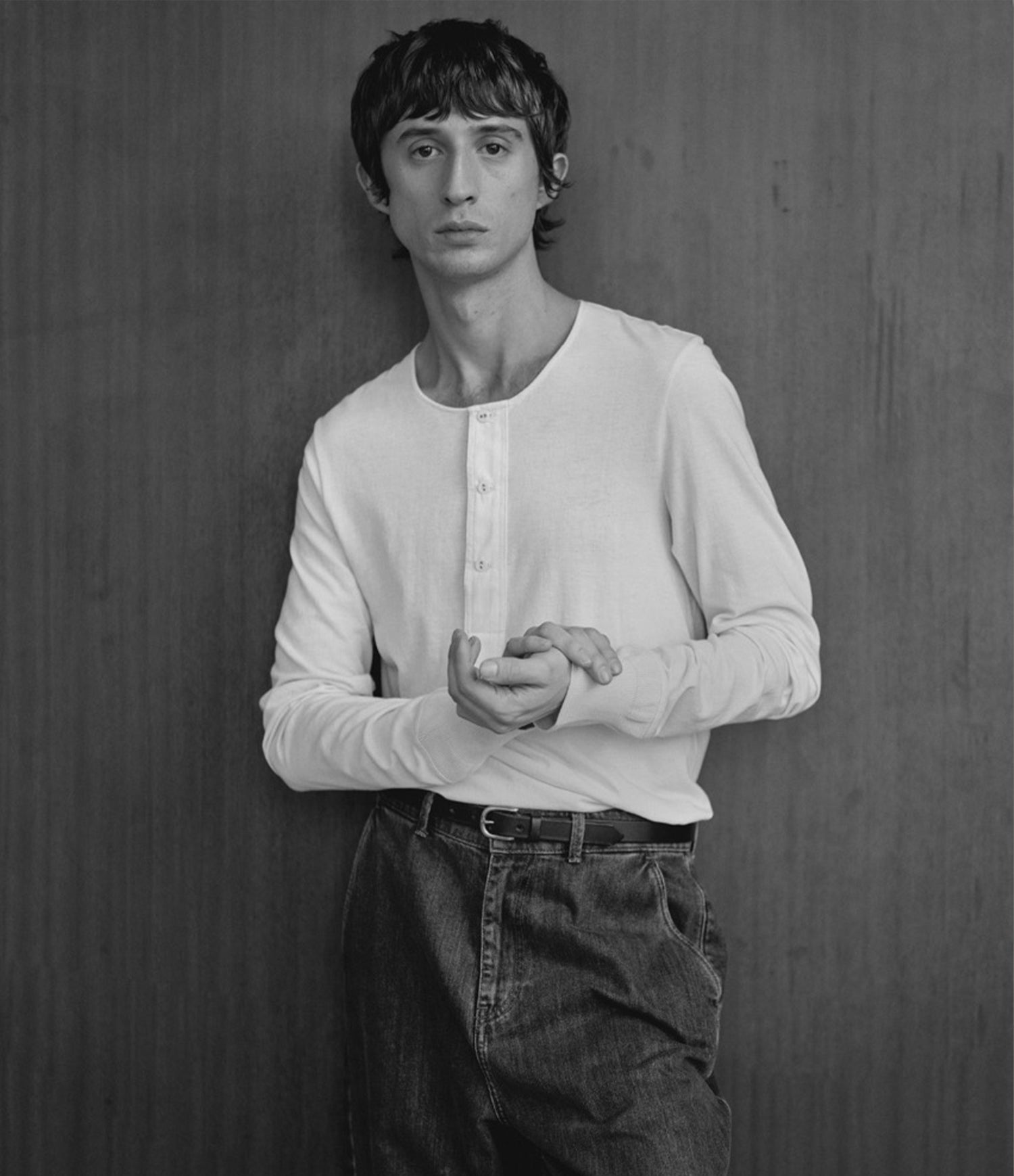 A black and white portrait of a young man wearing a white loopwheeled long-sleeve henley shirt and dark jeans, standing against a dark wood-paneled wall.