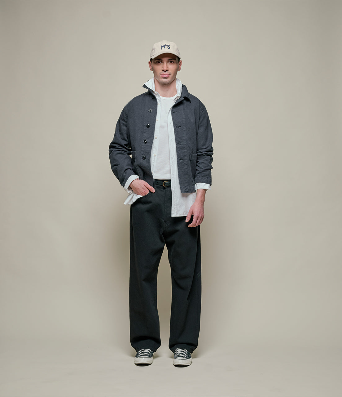 Person wearing a gray jacket, white shirt, black pants, and a cap on a beige background