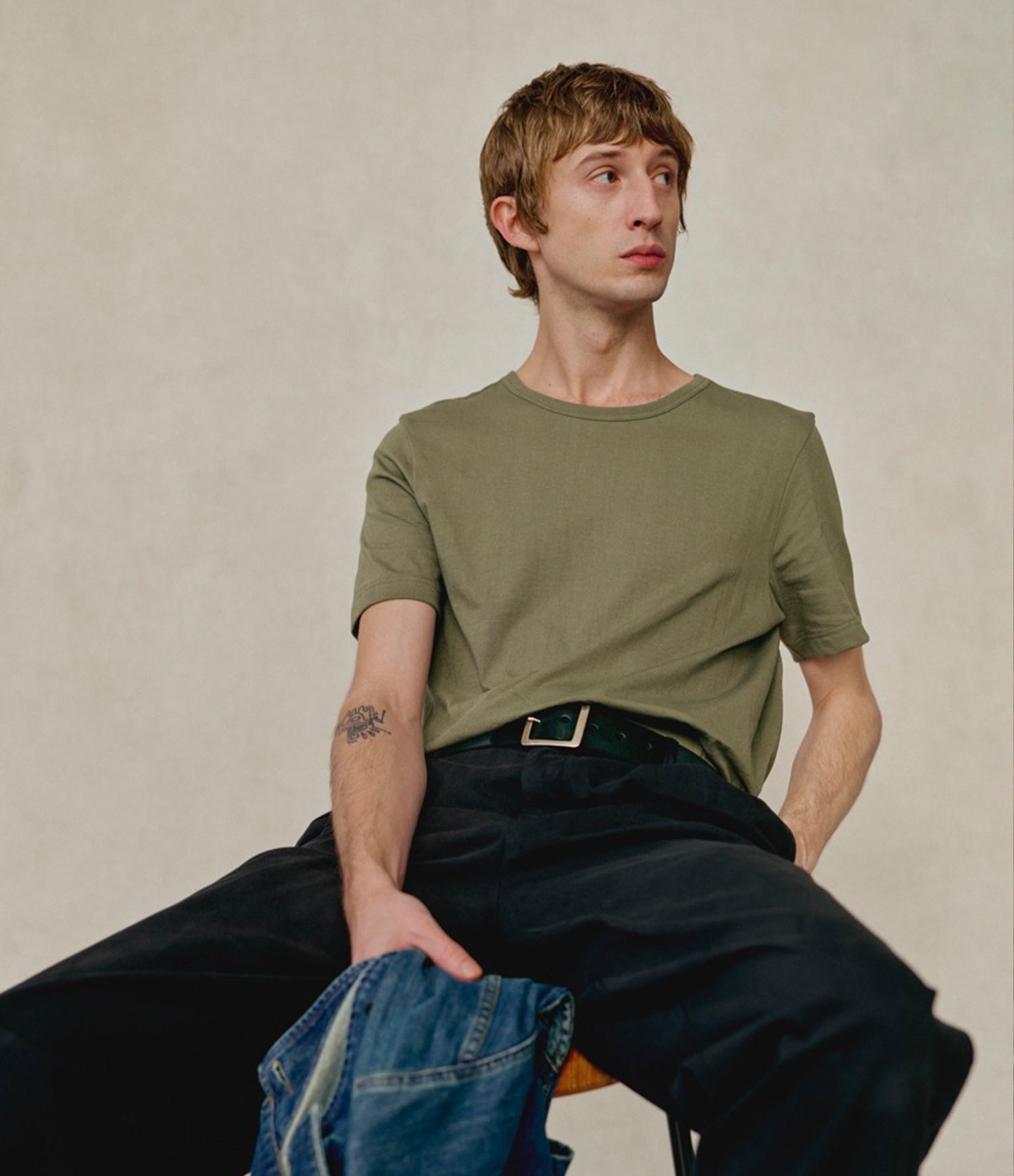 A man wearing an olive green loopwheeled short-sleeve crewneck t-shirt, dark wide-leg pants, and a green belt, sitting against a textured beige wall while holding a pair of blue jeans.