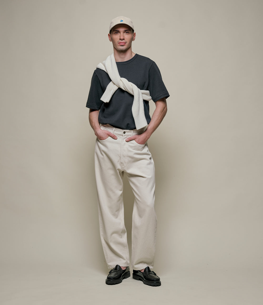 Man wearing a dark t-shirt, light pants, and a cap with a white towel over his shoulder against a beige background