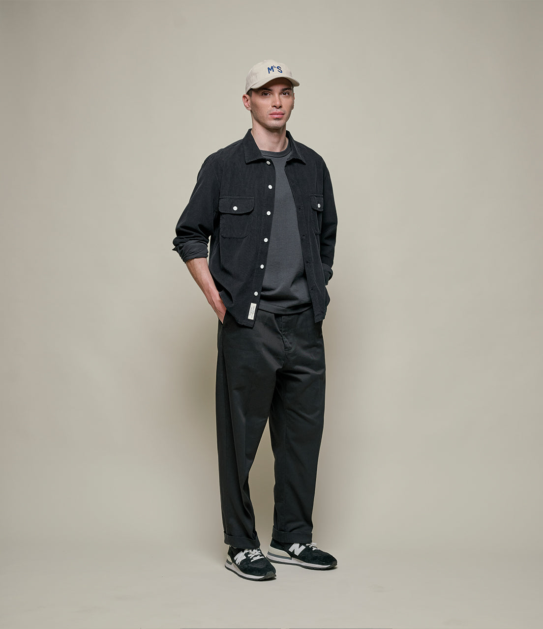 Man wearing a black jacket, gray shirt, black pants, and white cap on a beige background