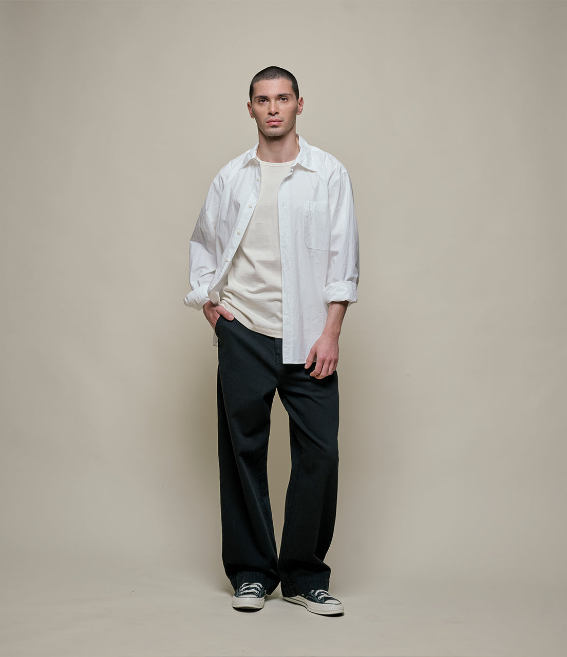 Man wearing a white shirt over a beige sweater with black pants against a plain background