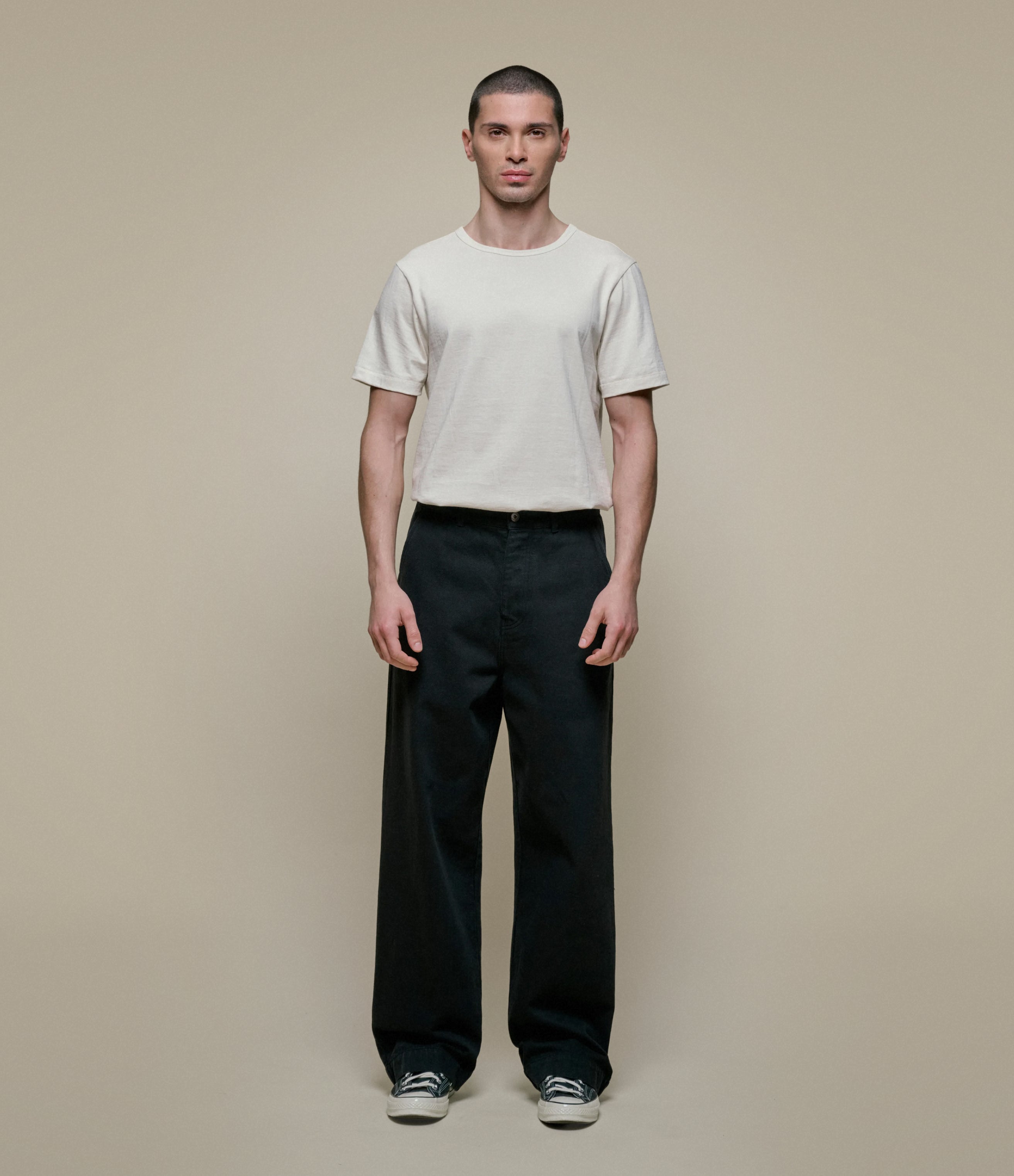 Man wearing a white shirt over a beige sweater with black pants against a plain background