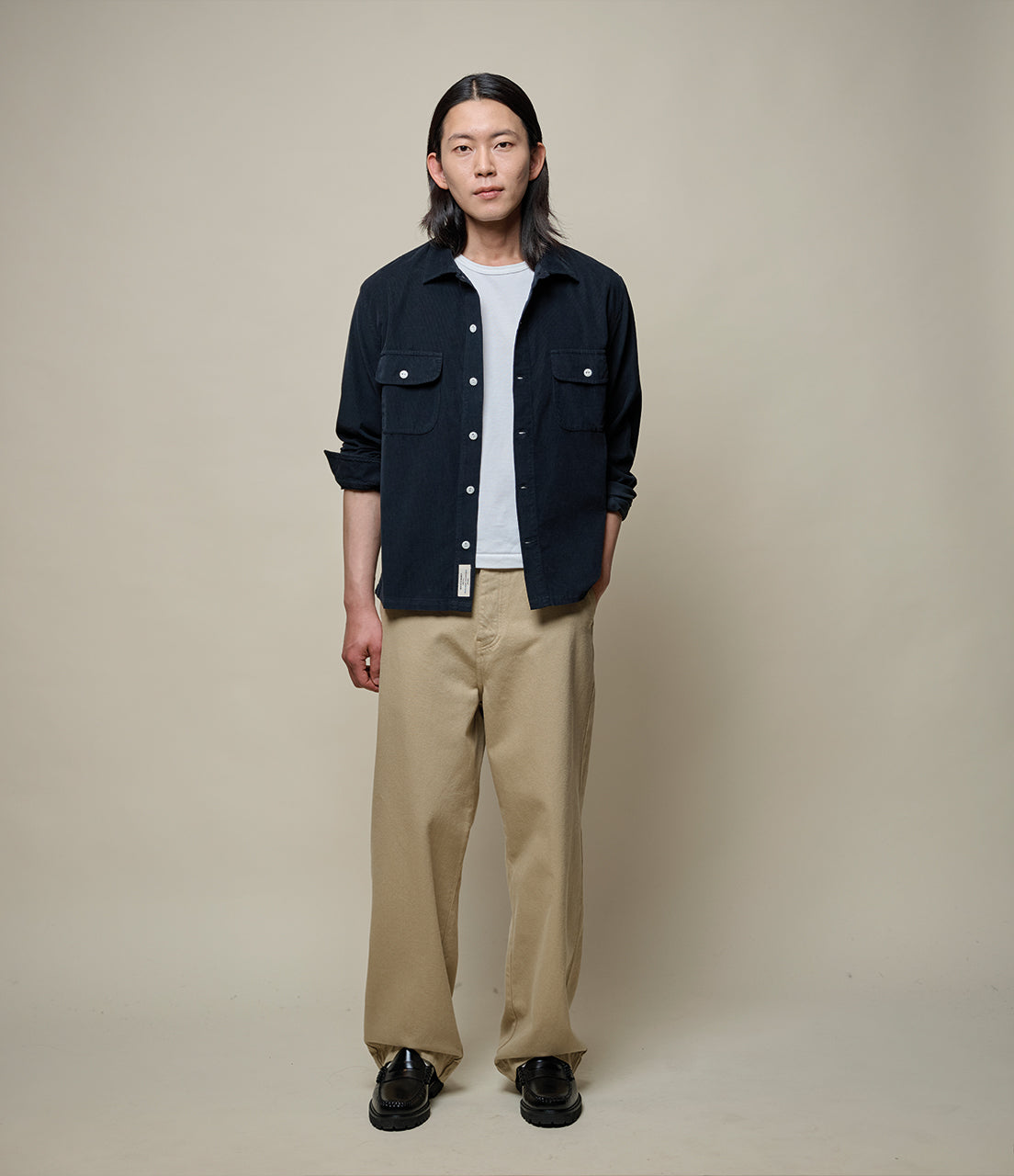 Person wearing a black corduroy shirt, white shirt, and beige pants against a plain background