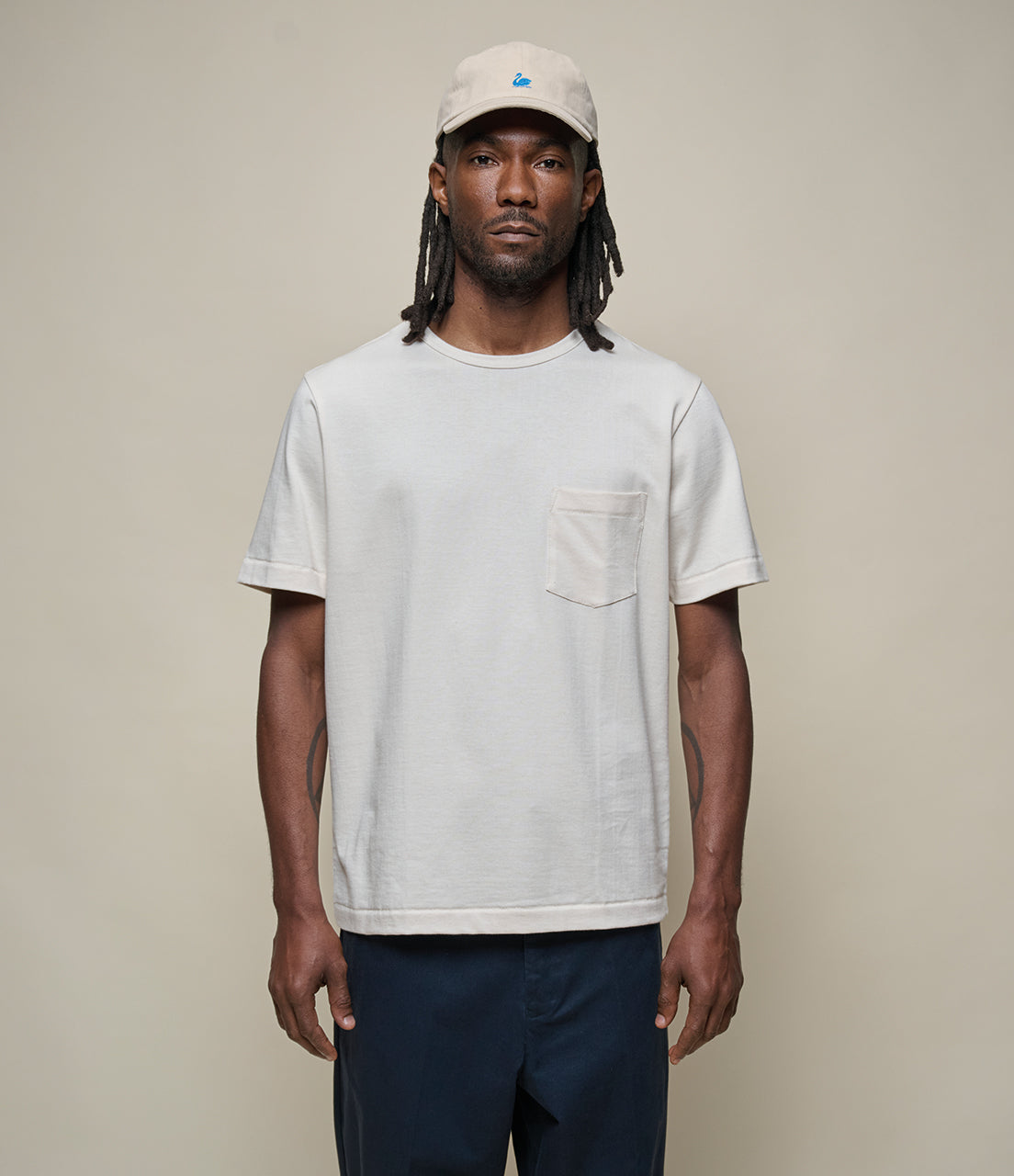 Man wearing a beige pocket t-shirt and beige cap against a plain background