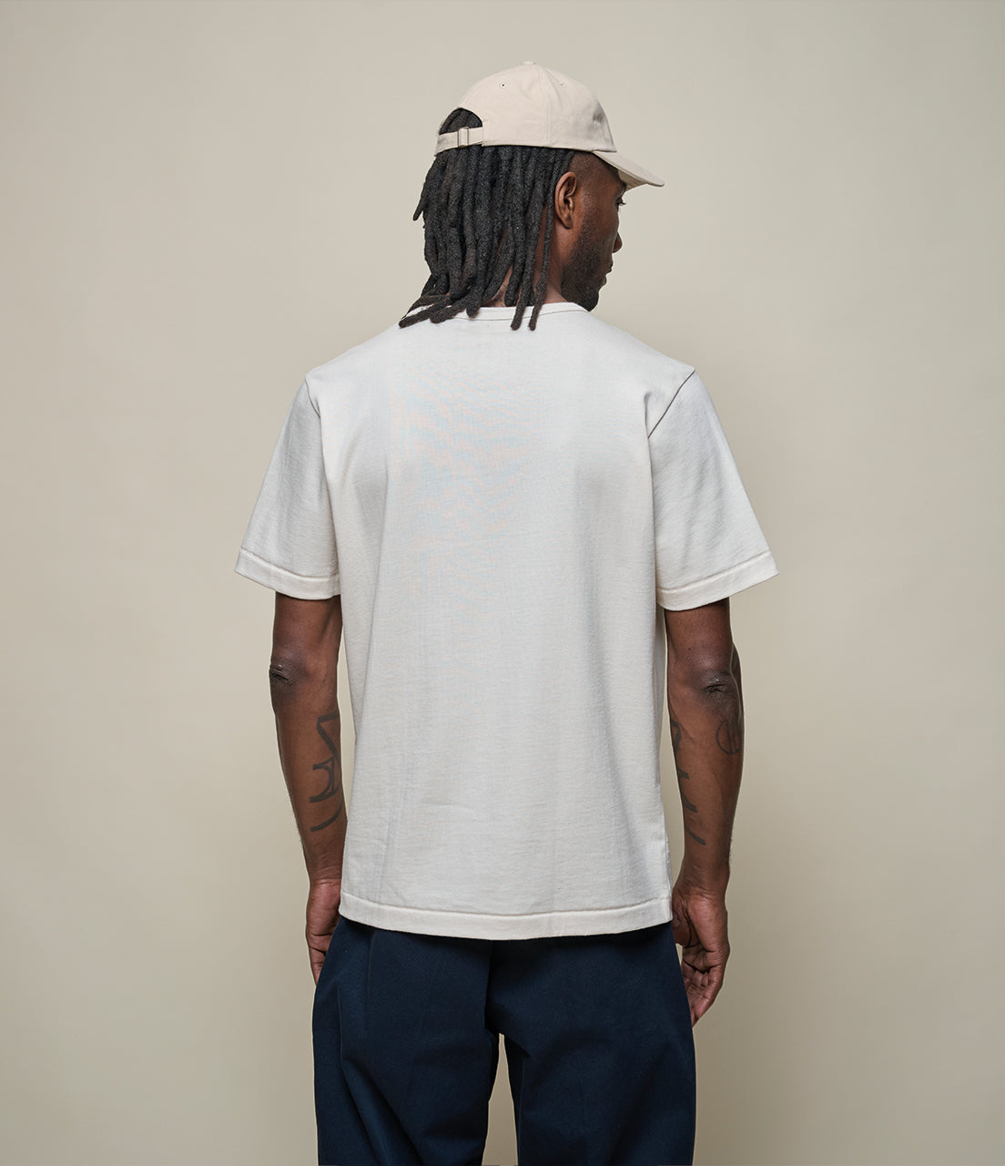 Person wearing a beige t-shirt and navy pants with a beige cap, standing against a plain background