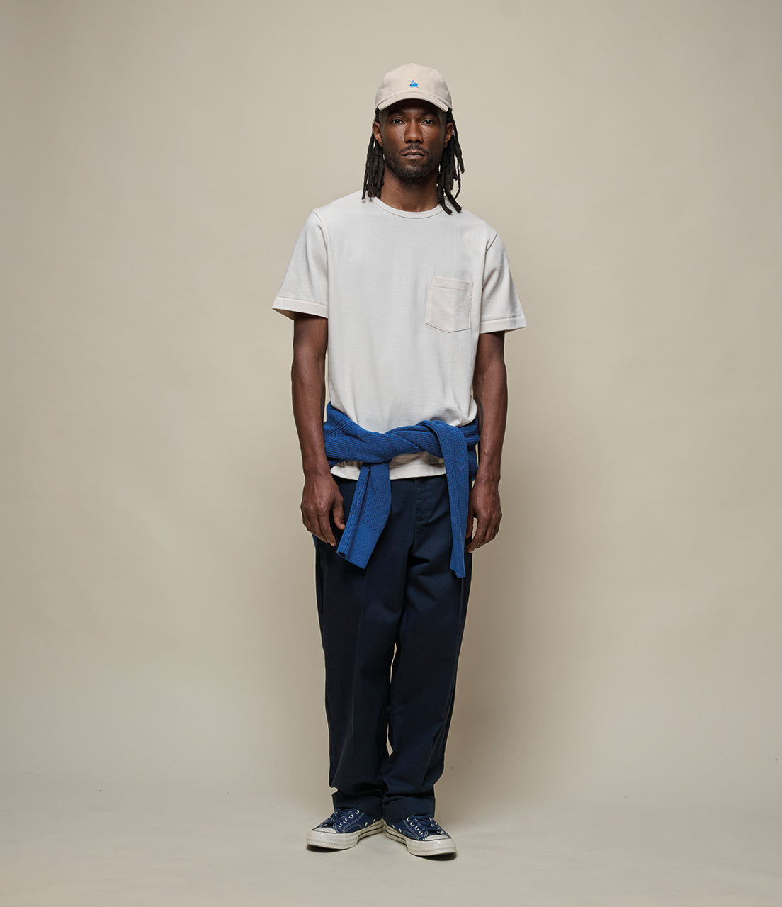 Man wearing a beige t-shirt, blue jacket, dark pants, and a cap on a beige background