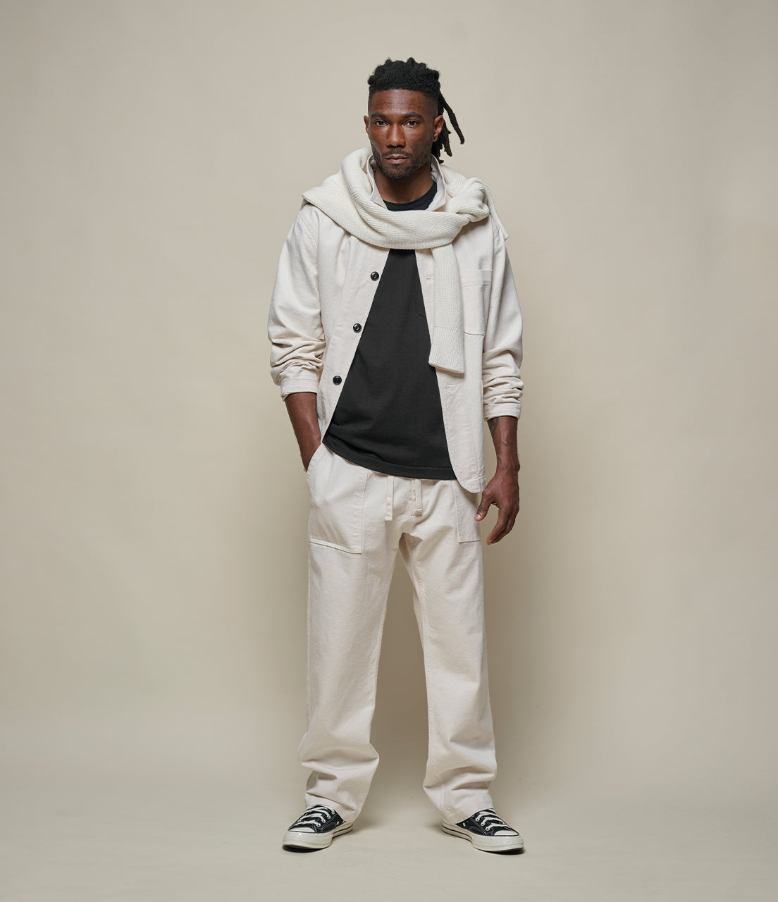 Man wearing a white hoodie, black shirt, beige pants, and black shoes against a plain background