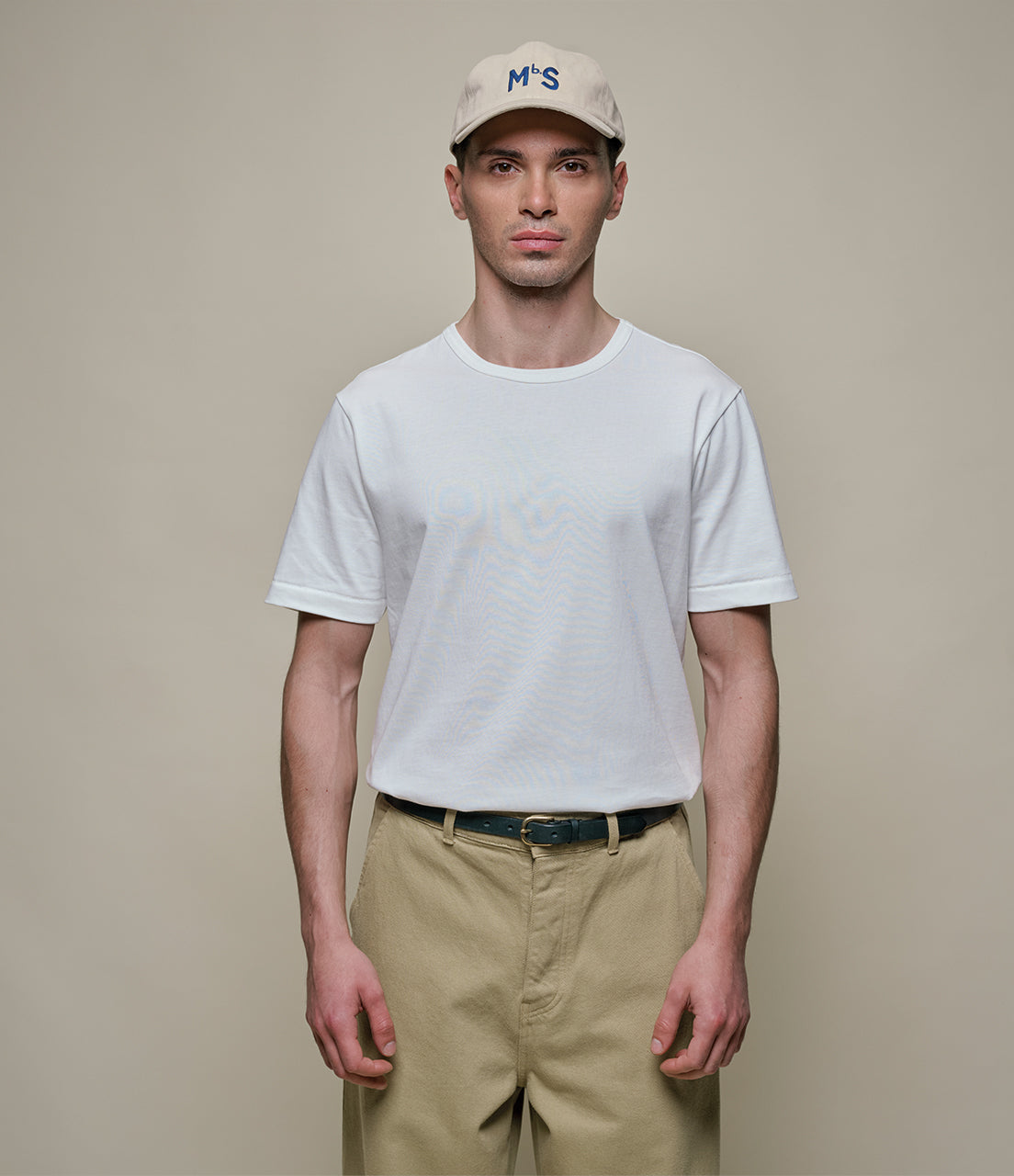 Man wearing a white t-shirt, beige cap, and khaki pants against a plain background