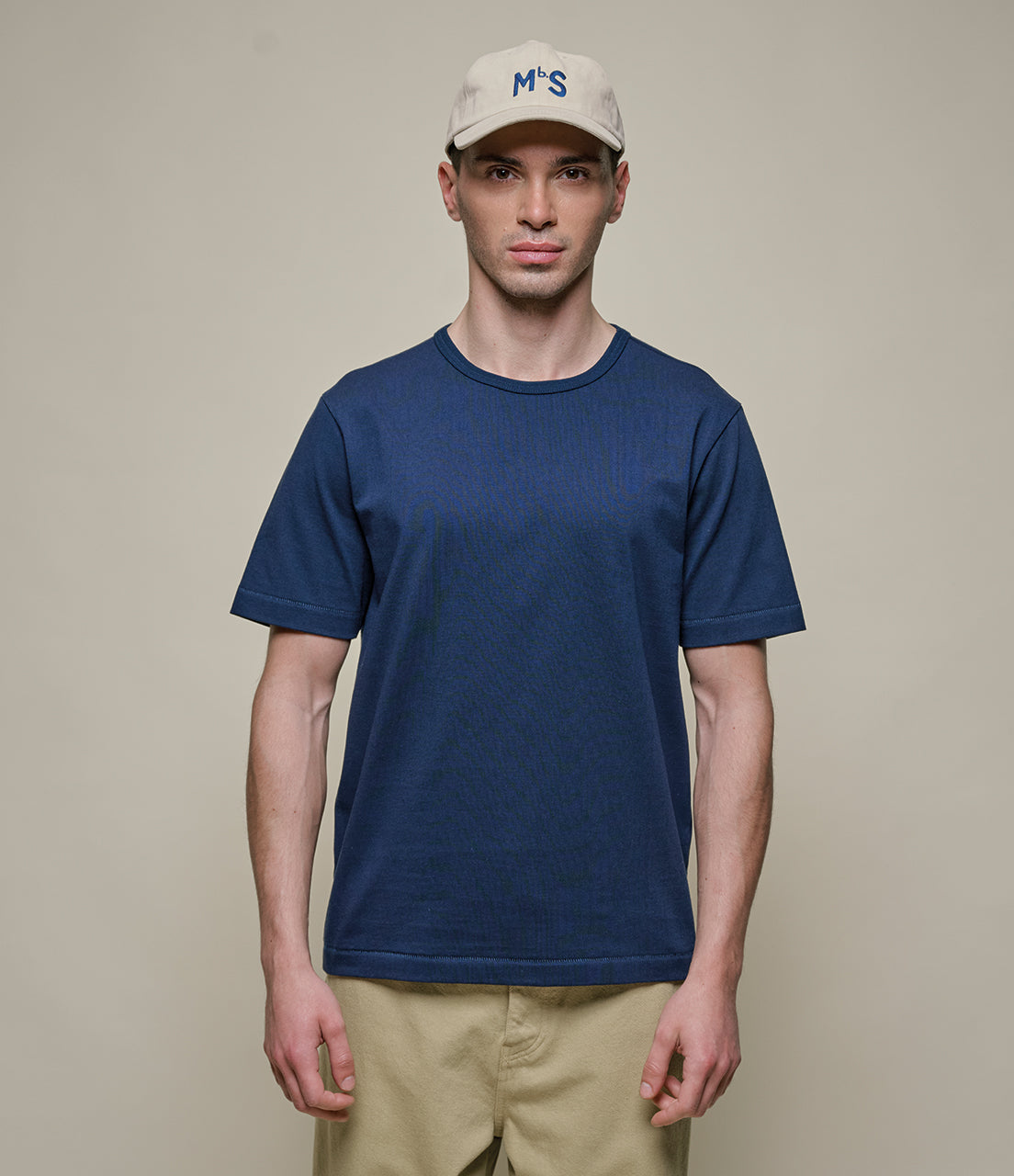 Man wearing a blue t-shirt and beige cap against a plain background