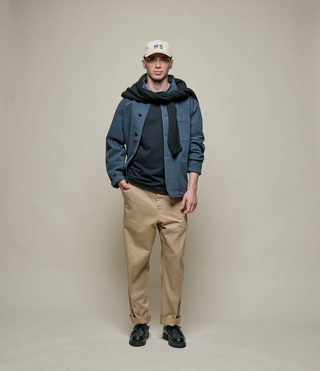 Person wearing a blue jacket, black hoodie, beige pants, and a white cap on a plain background