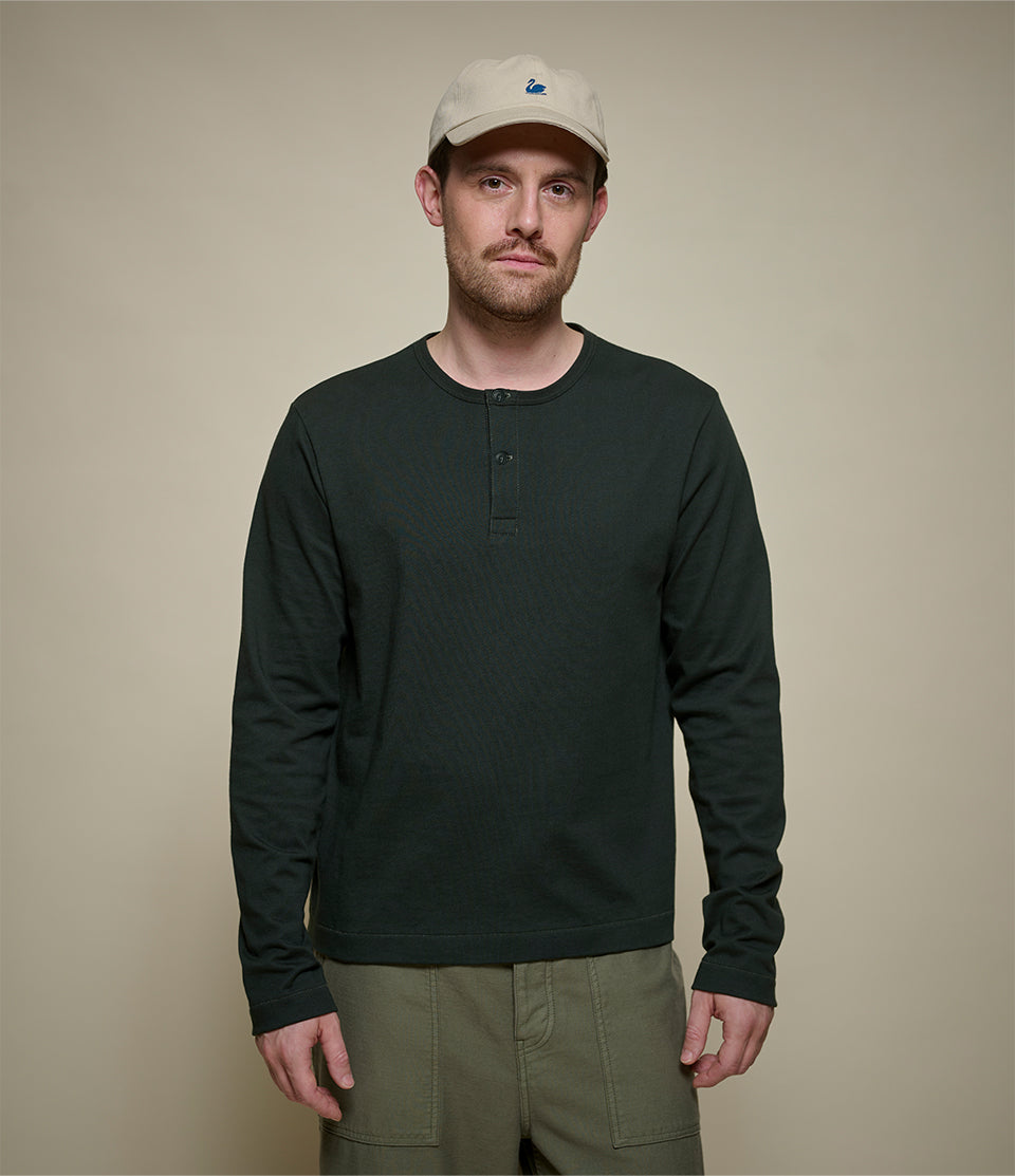 A man wearing a dark forest green cropped long-sleeve henley shirt, olive green cargo pants, and a beige baseball cap, standing against a solid beige background.