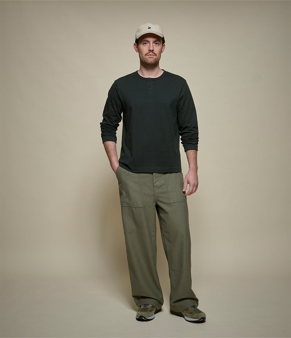 A man wearing a dark green cropped long-sleeve henley shirt, olive green cargo pants, and a beige baseball cap, standing against a solid beige studio background.
