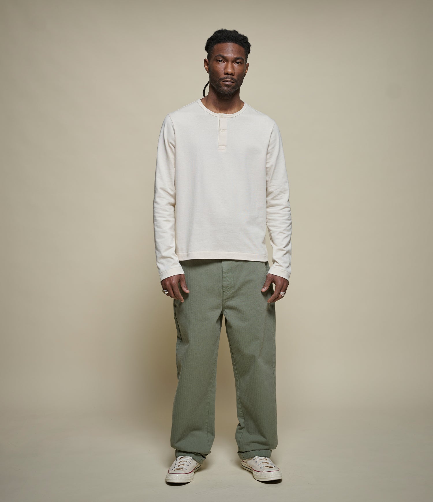 A man modeling a beige cropped long-sleeve henley shirt and olive green pants against a solid beige studio background.