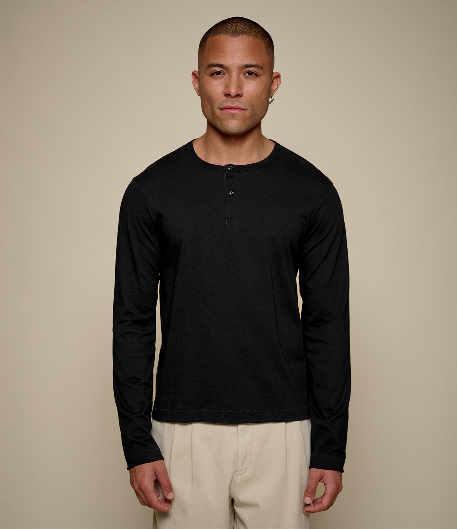 A man wearing a black cropped long-sleeve henley shirt and beige pants, standing against a solid beige background.