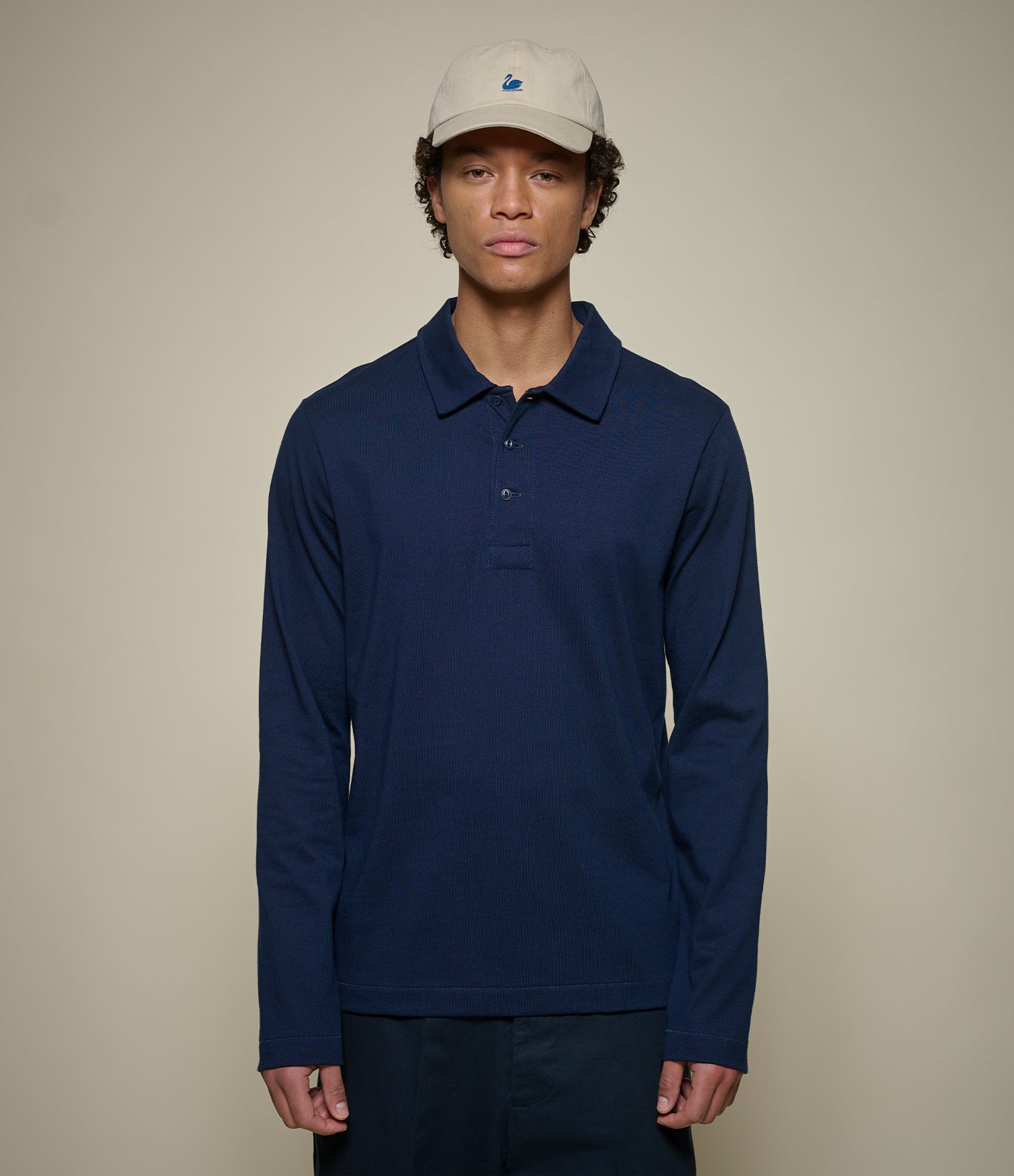 A man wearing a navy blue long-sleeve polo shirt and a light beige baseball cap, standing against a plain beige studio background.