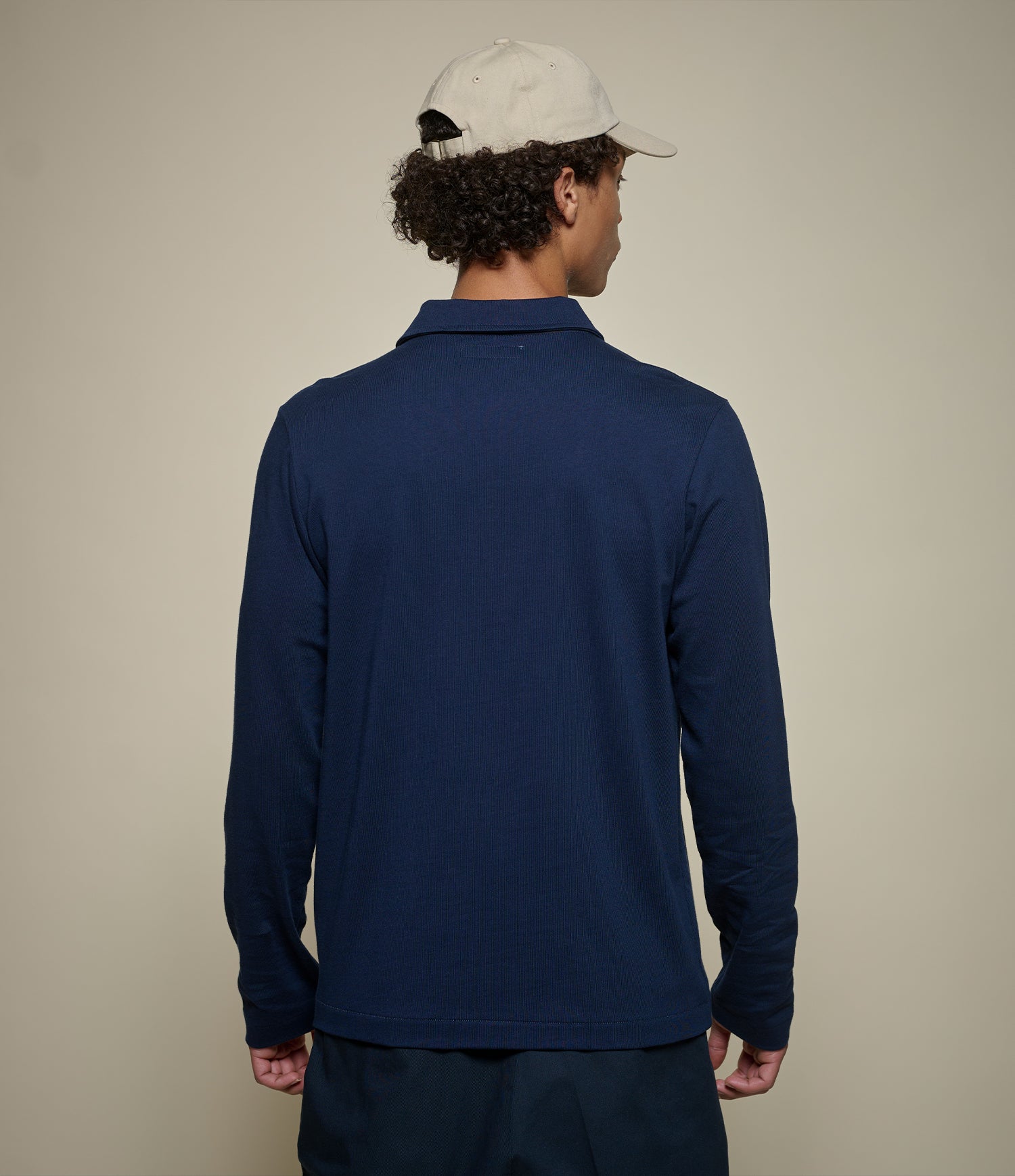 A man viewed from behind wearing a navy blue long-sleeve polo shirt, a light beige baseball cap, and dark pants, standing against a plain beige studio background.