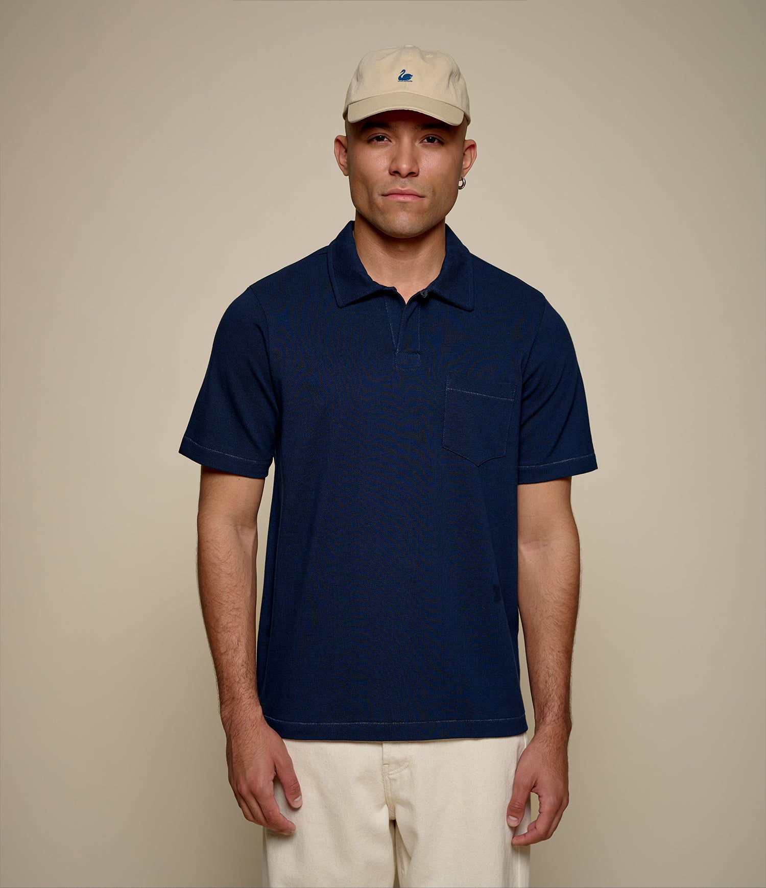 Man wearing a navy blue loopwheeled pikée polo shirt and beige cap against a beige background