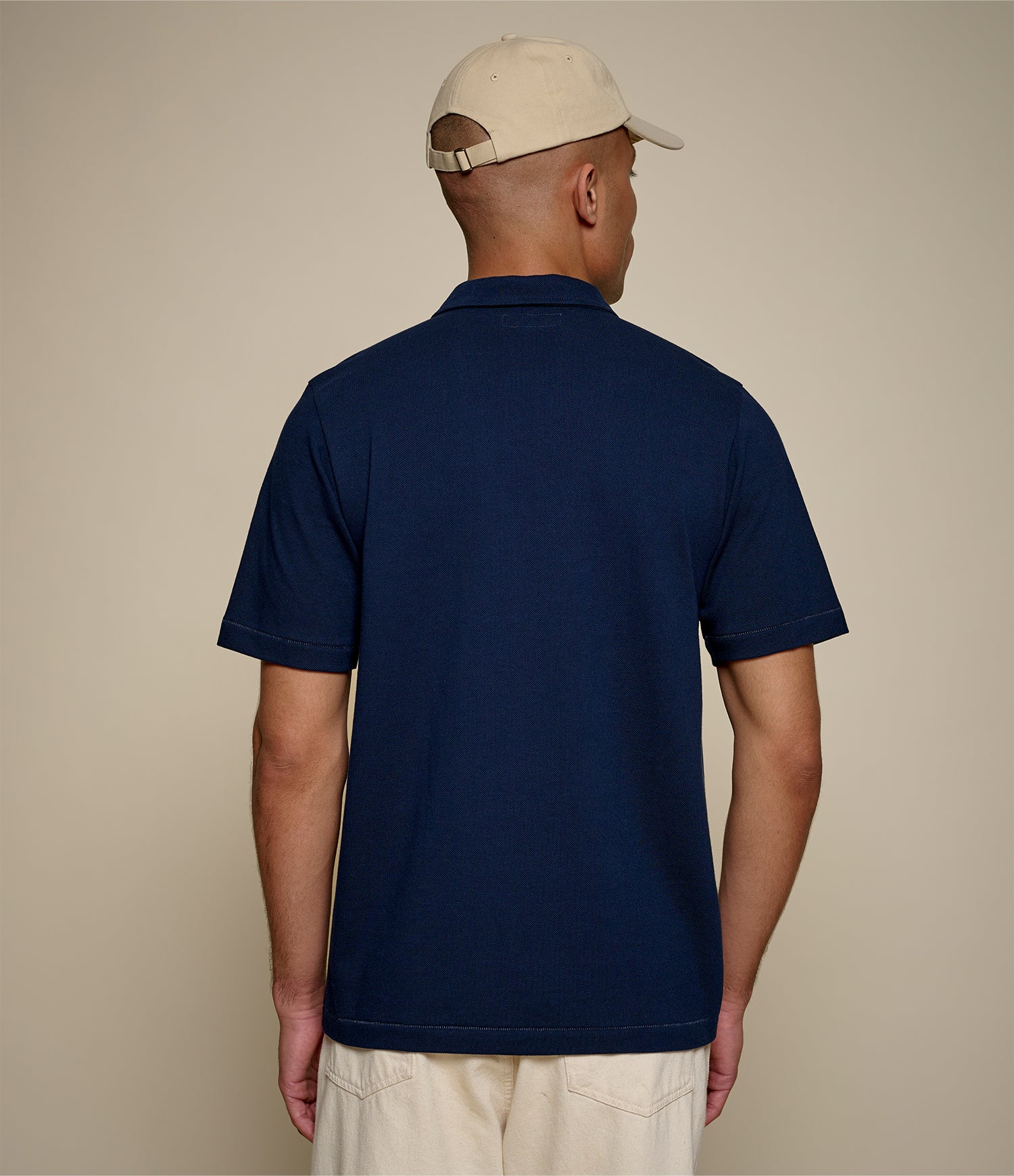 Man wearing a navy blue loopwheeled pikée polo shirt and beige cap on a beige background